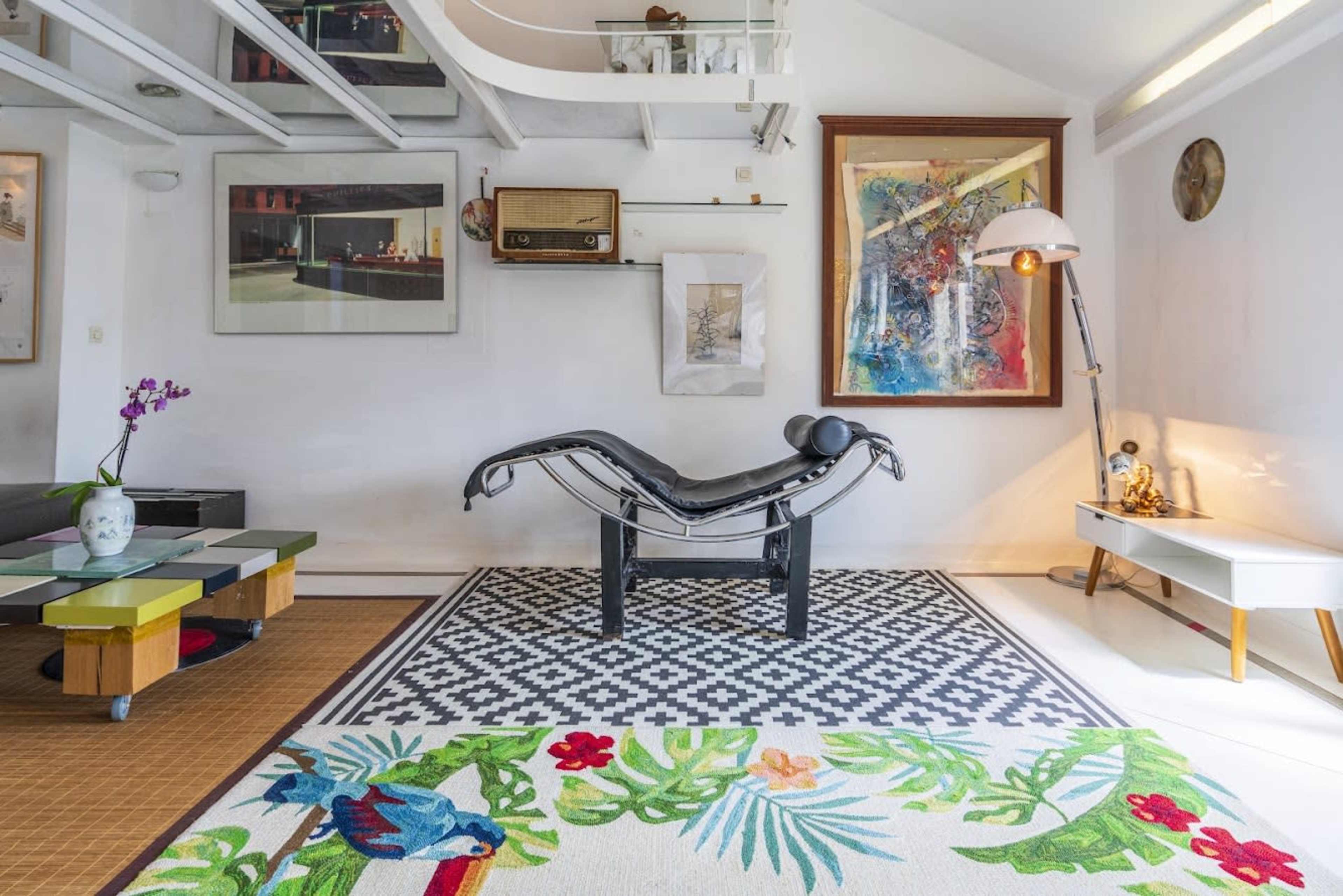 The image shows a modern interior featuring a unique chaise lounge chair, a colorful area rug, and various decorative art pieces on the walls.