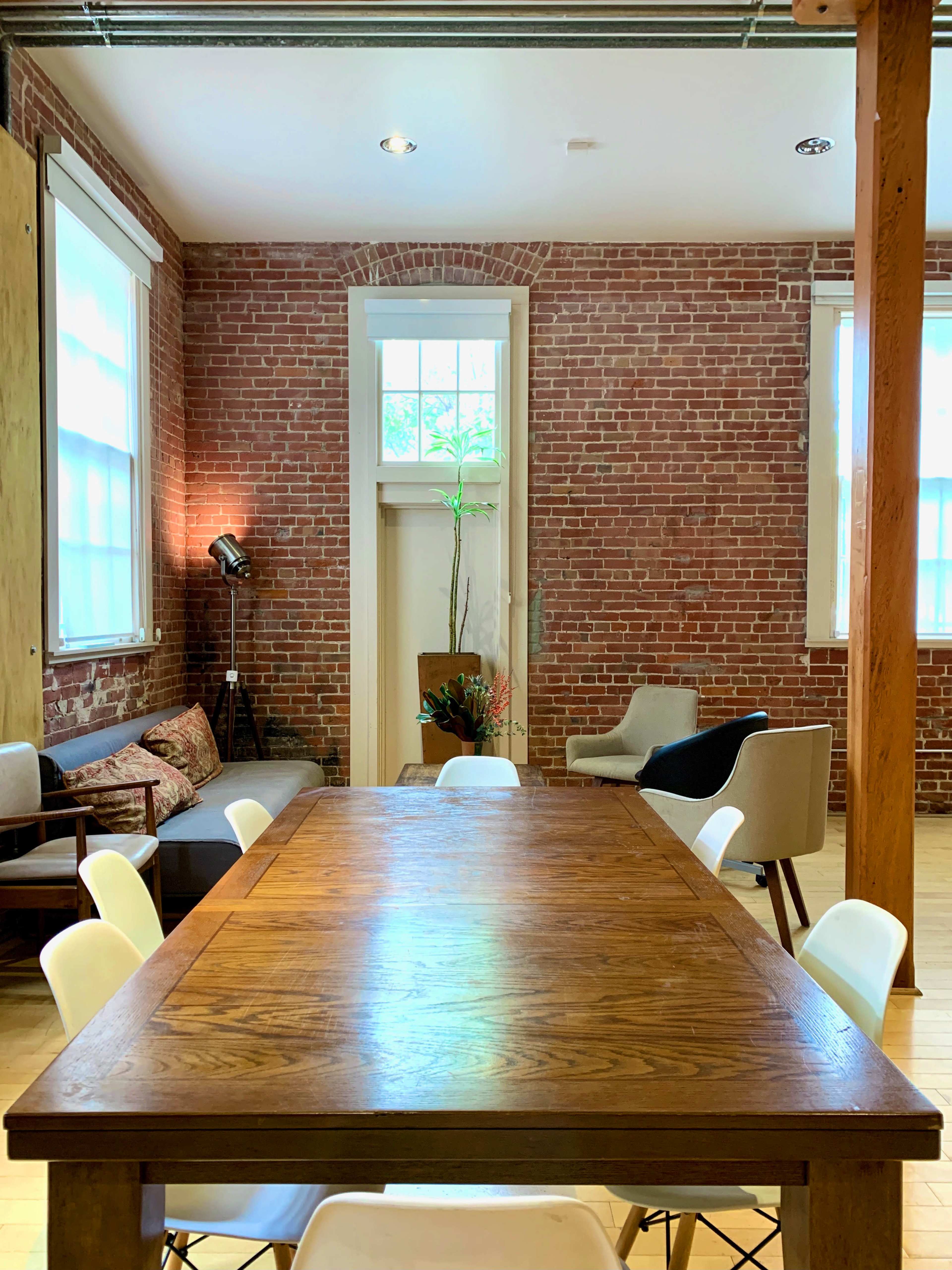 A spacious room with exposed brick walls, a large wooden table in the center, and various chairs and seating arrangements along the sides.