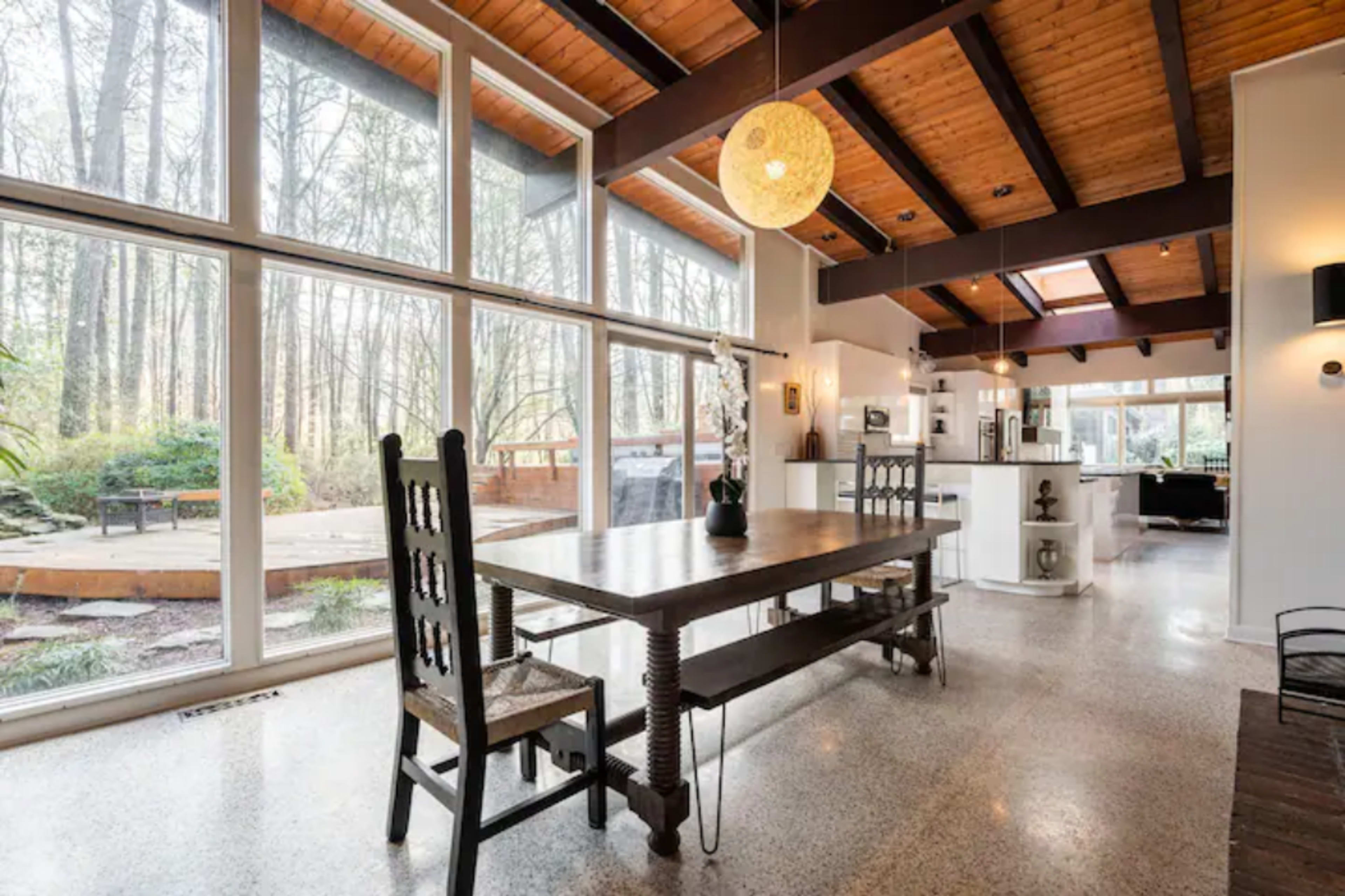 The image shows a dining area with a wooden table and chairs, large windows overlooking a wooded view, and a modern kitchen in the background.