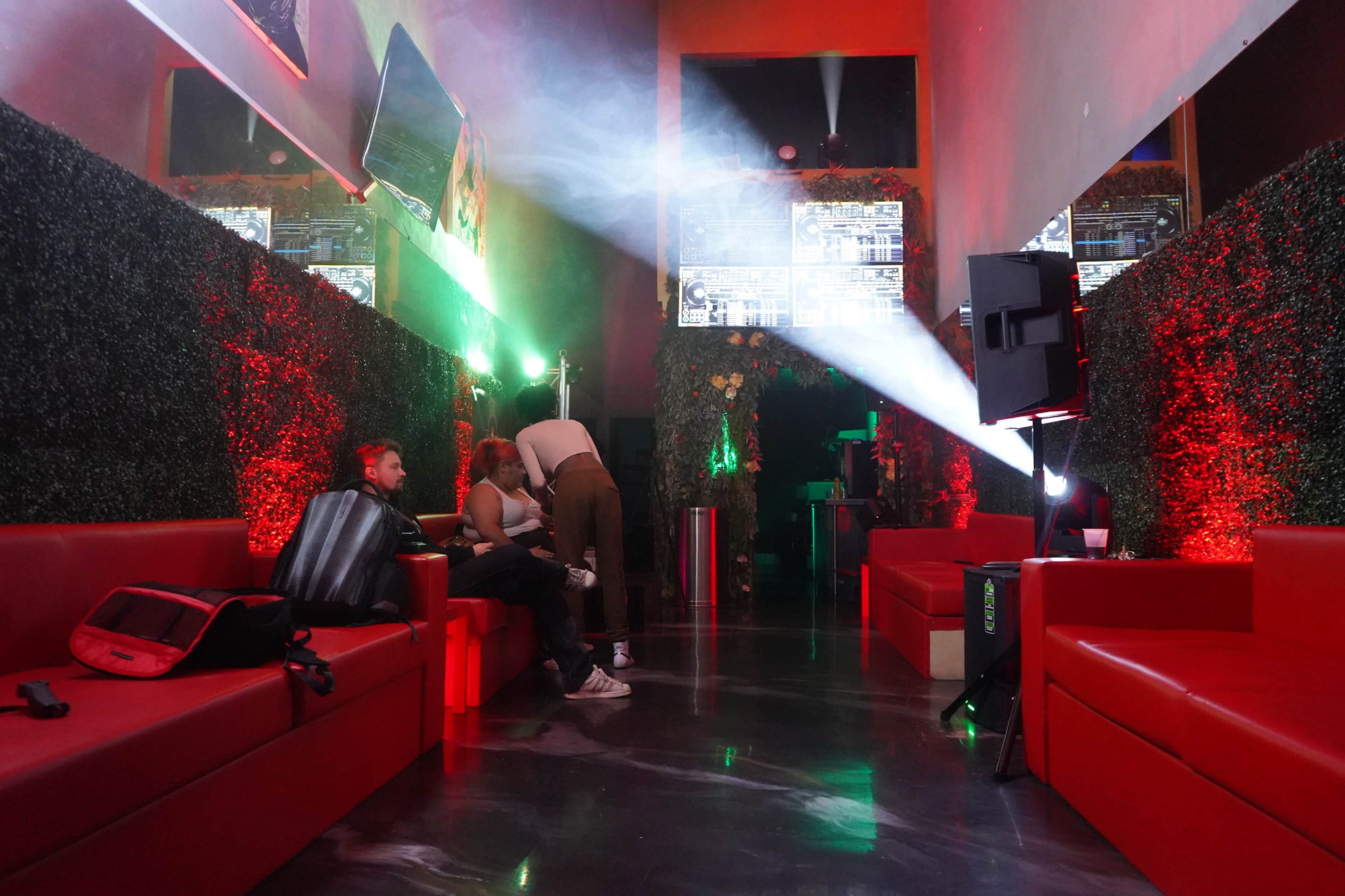 The image shows a dimly lit lounge area with red couches, greenery on the walls, and a DJ setup at the back, illuminated by colorful lights and projections.