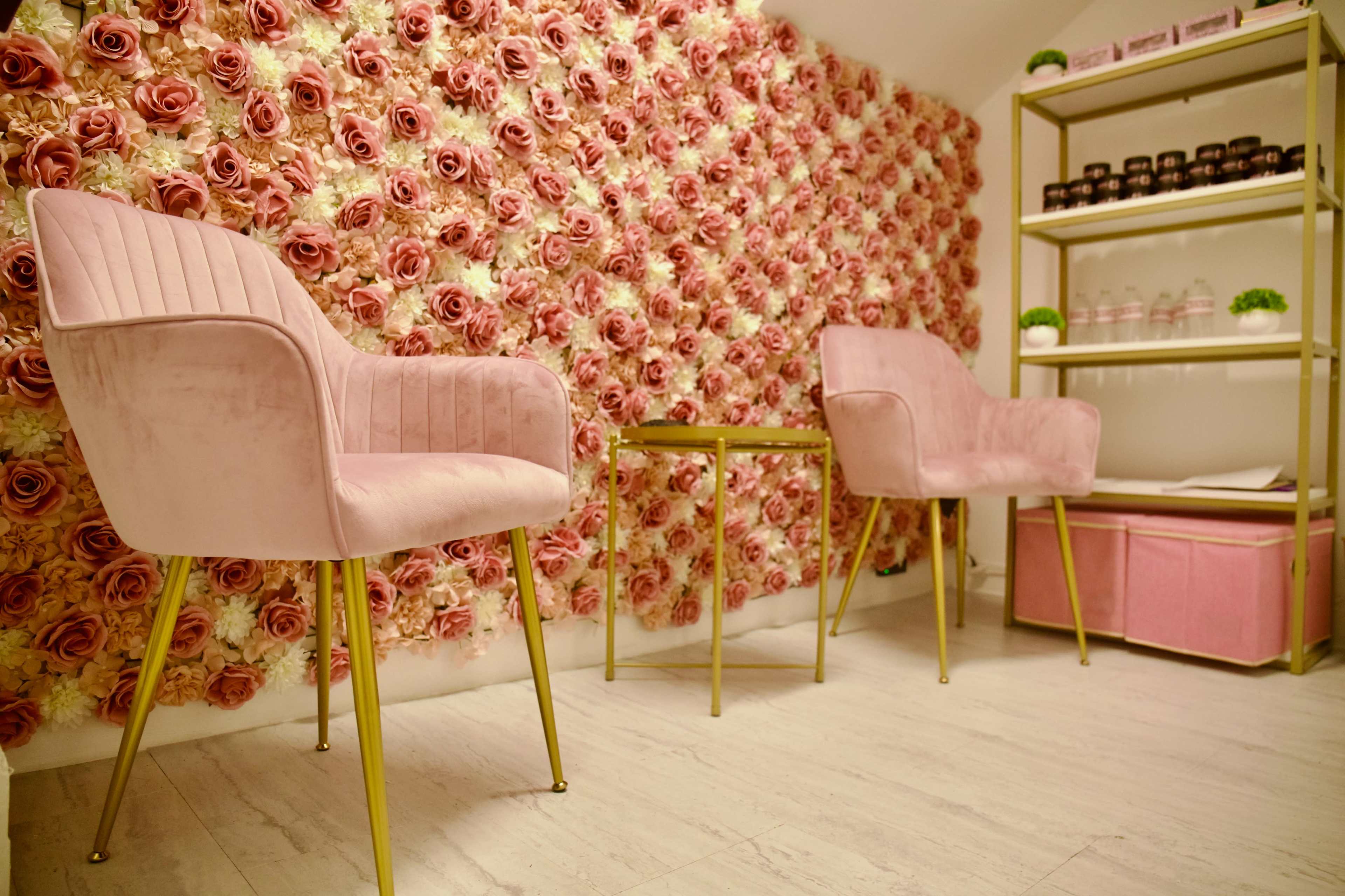 The image shows two pink velvet chairs positioned at a small gold table in front of a wall adorned with artificial roses, along with a shelf displaying jars and decorative items.