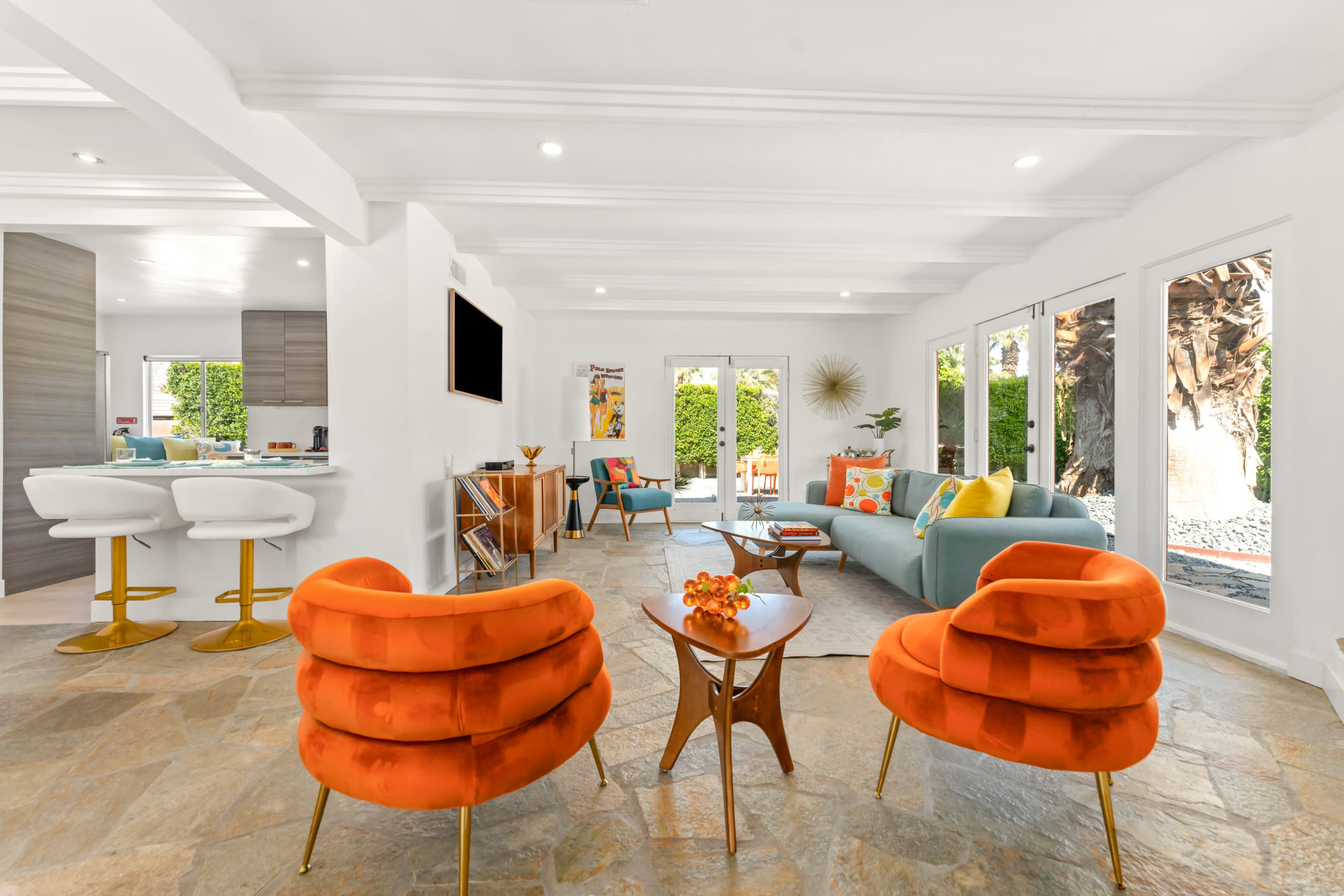 A bright living room features orange chairs, a blue sofa, a coffee table, and large windows overlooking a garden.