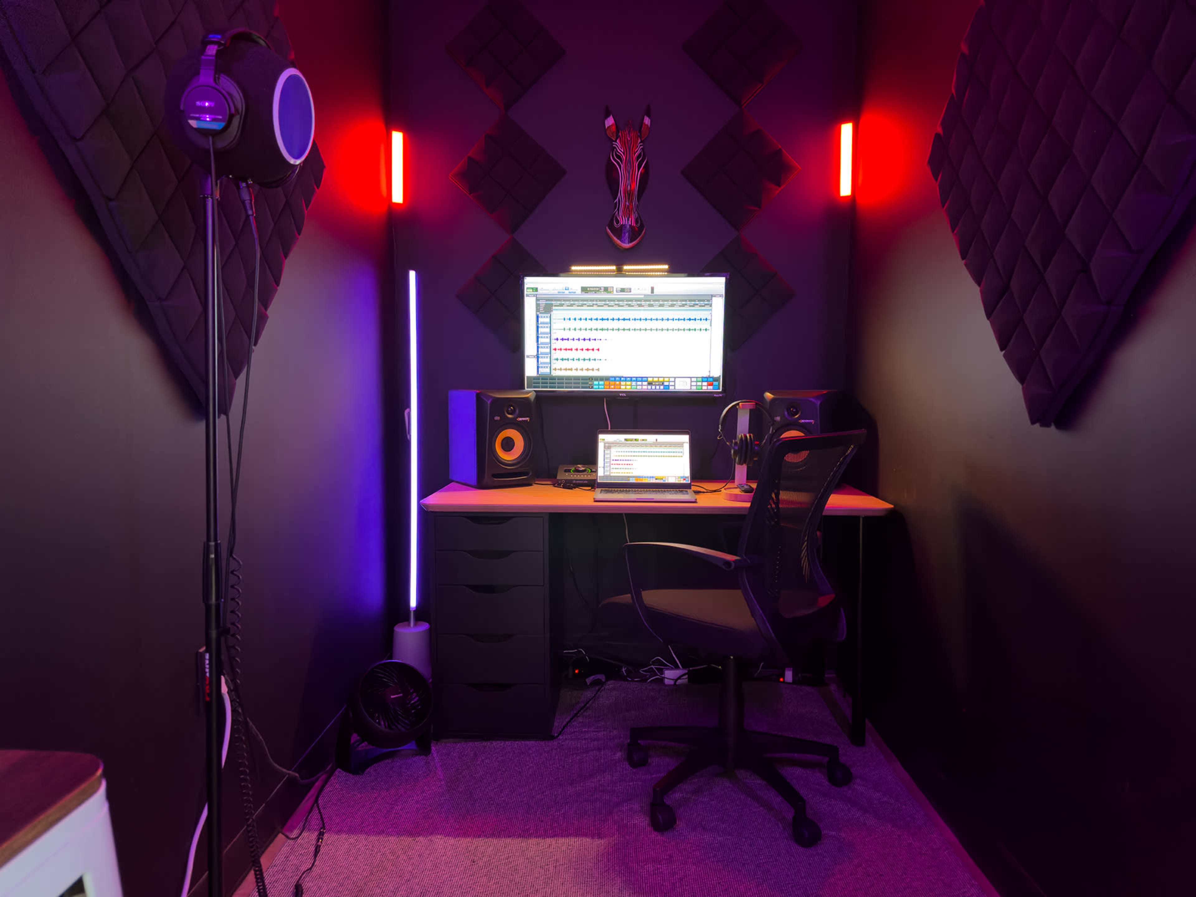 The image shows a recording studio with a computer workstation, speakers, and acoustic foam panels on the walls, illuminated by colorful LED lights.