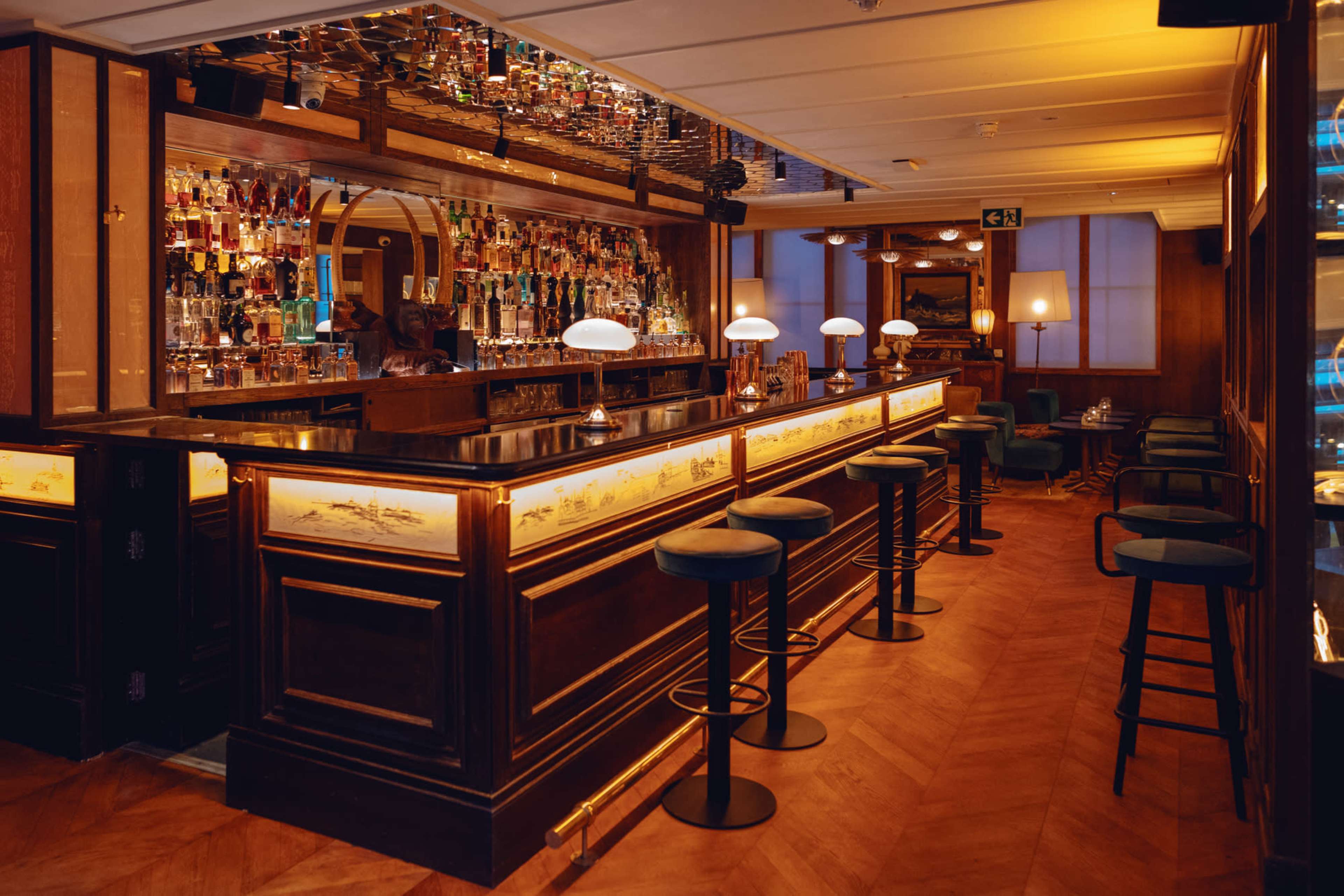 A dimly lit bar featuring a long wooden counter with high stools, lined with bottles of various spirits on the shelves behind it.