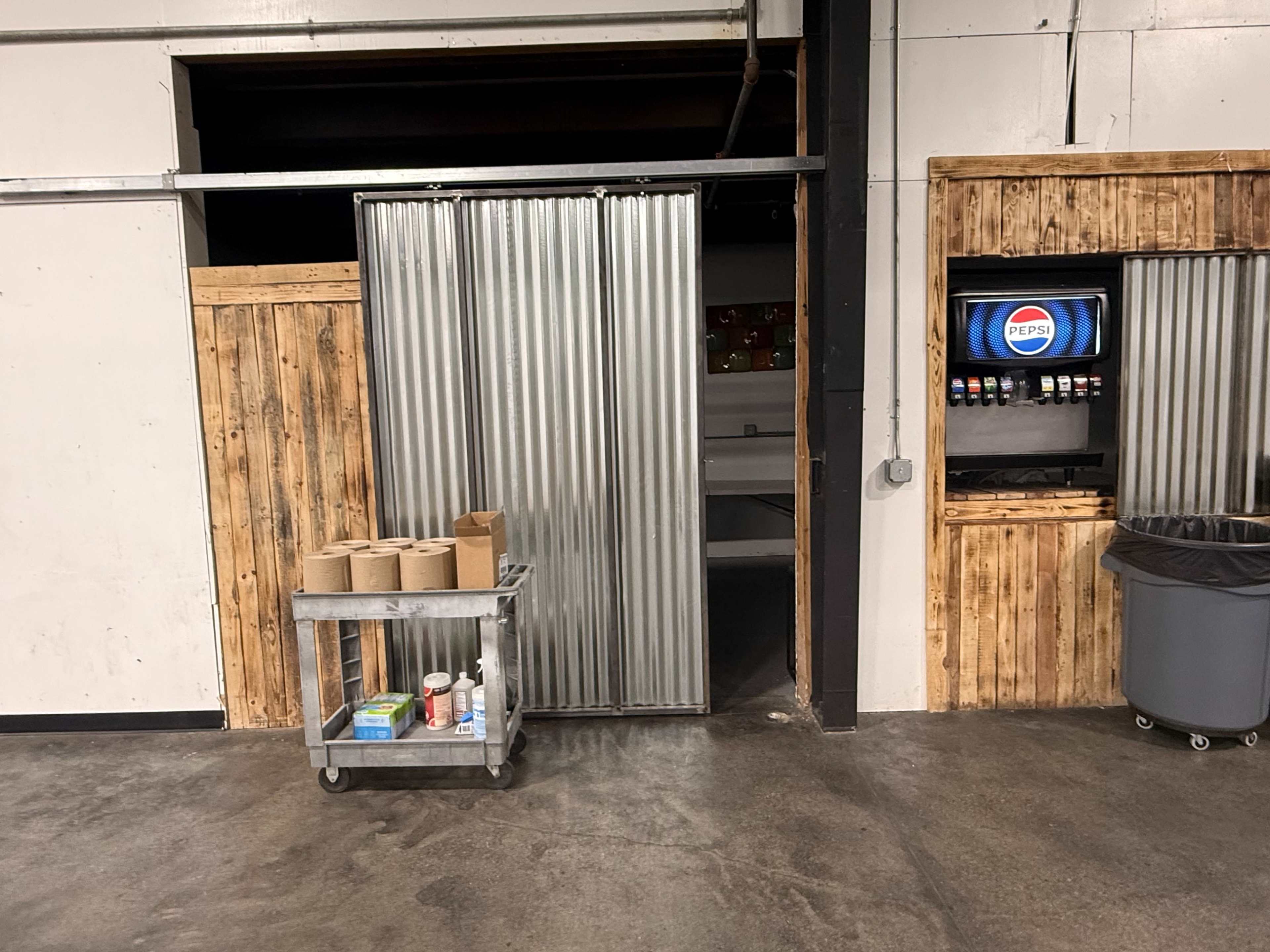 The image shows a metallic sliding door opening into a dimly lit room with a wooden cart holding various supplies in front of it, and a soda machine on the right.