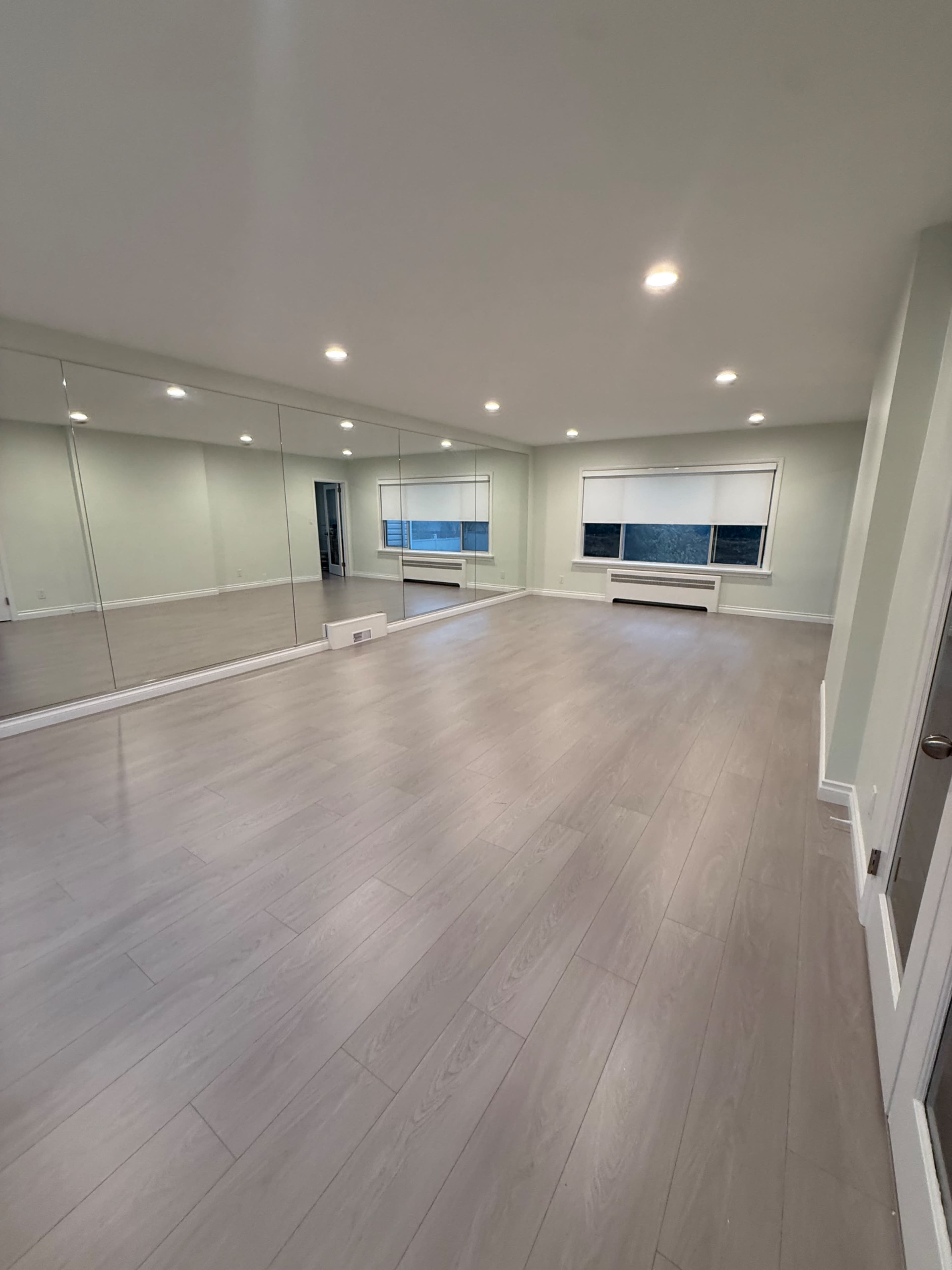 The image shows a spacious room with light gray flooring, mirrored wall panels, and large windows with blinds.