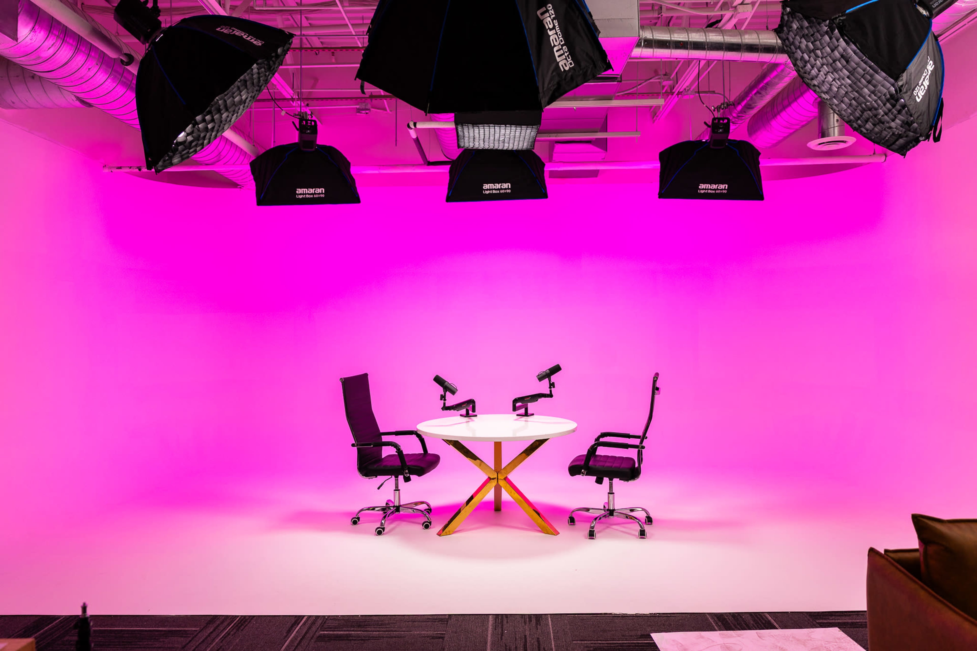 A brightly lit studio features two black chairs positioned at a white round table, set against a pink backdrop with multiple overhead lights.