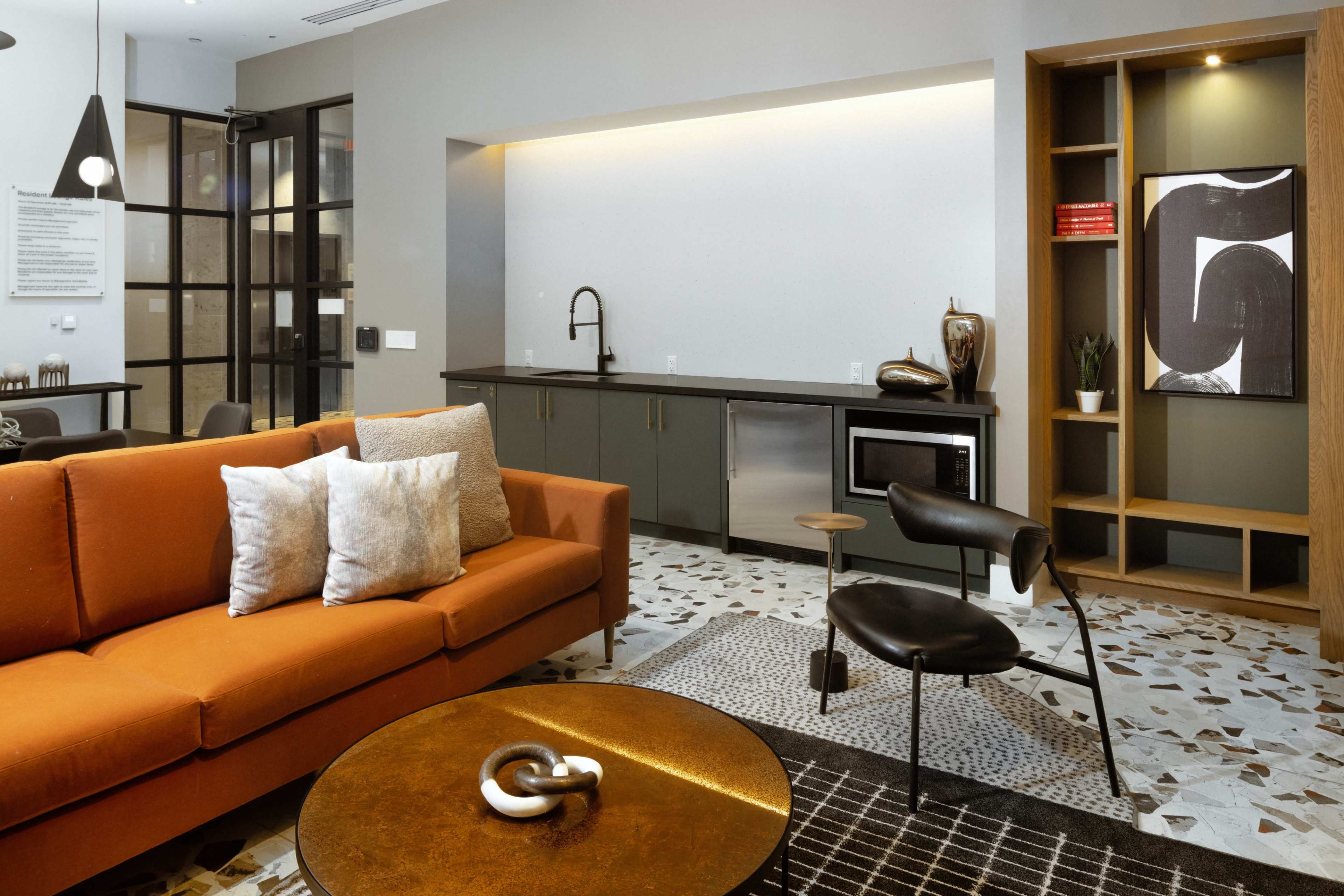 A modern living room with an orange couch, a round coffee table, and a sleek kitchenette area against a neutral-colored wall.