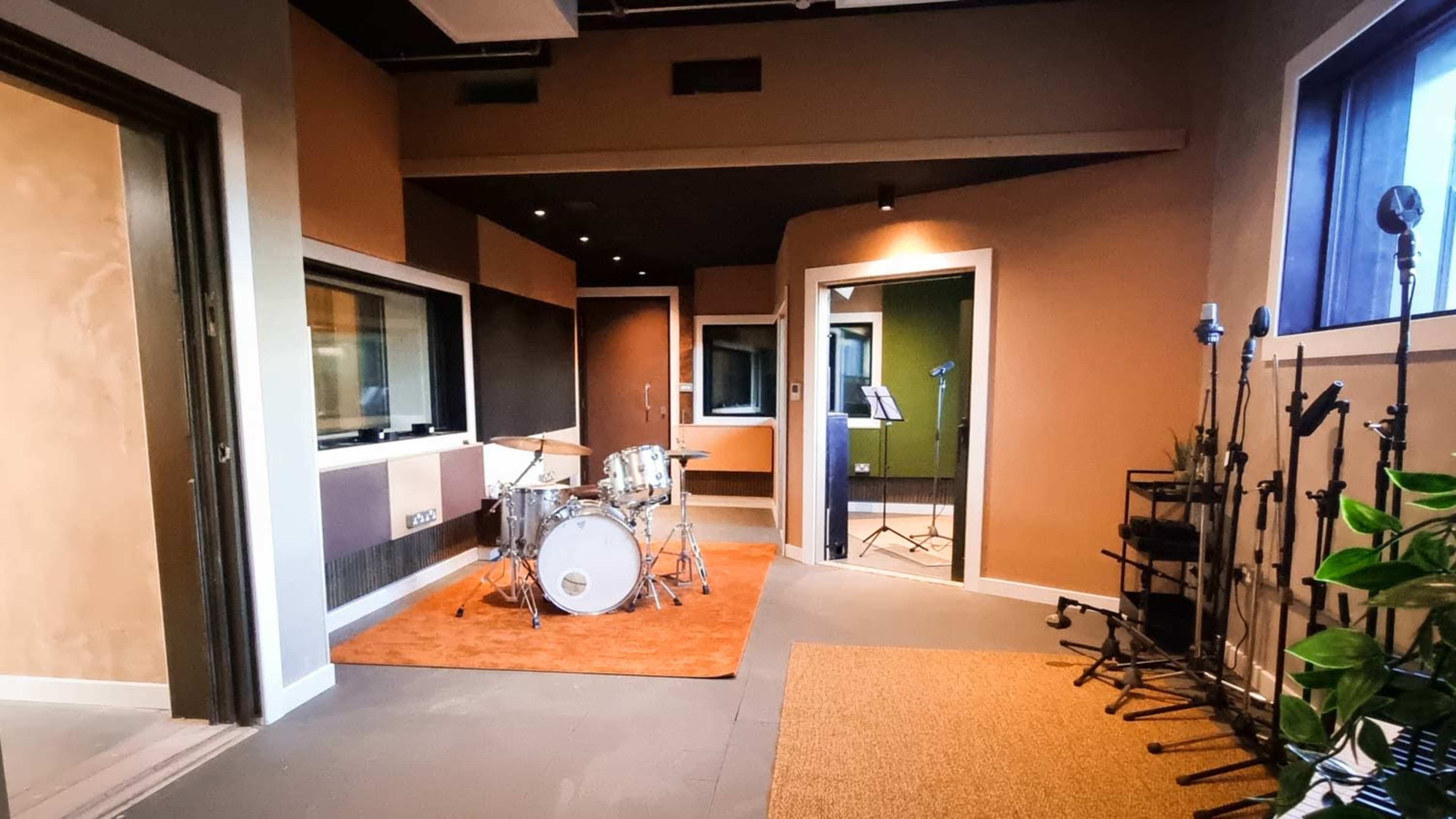 A soundproof recording studio with a drum set in one corner, various microphones positioned around the room, and a glass window separating different sections.