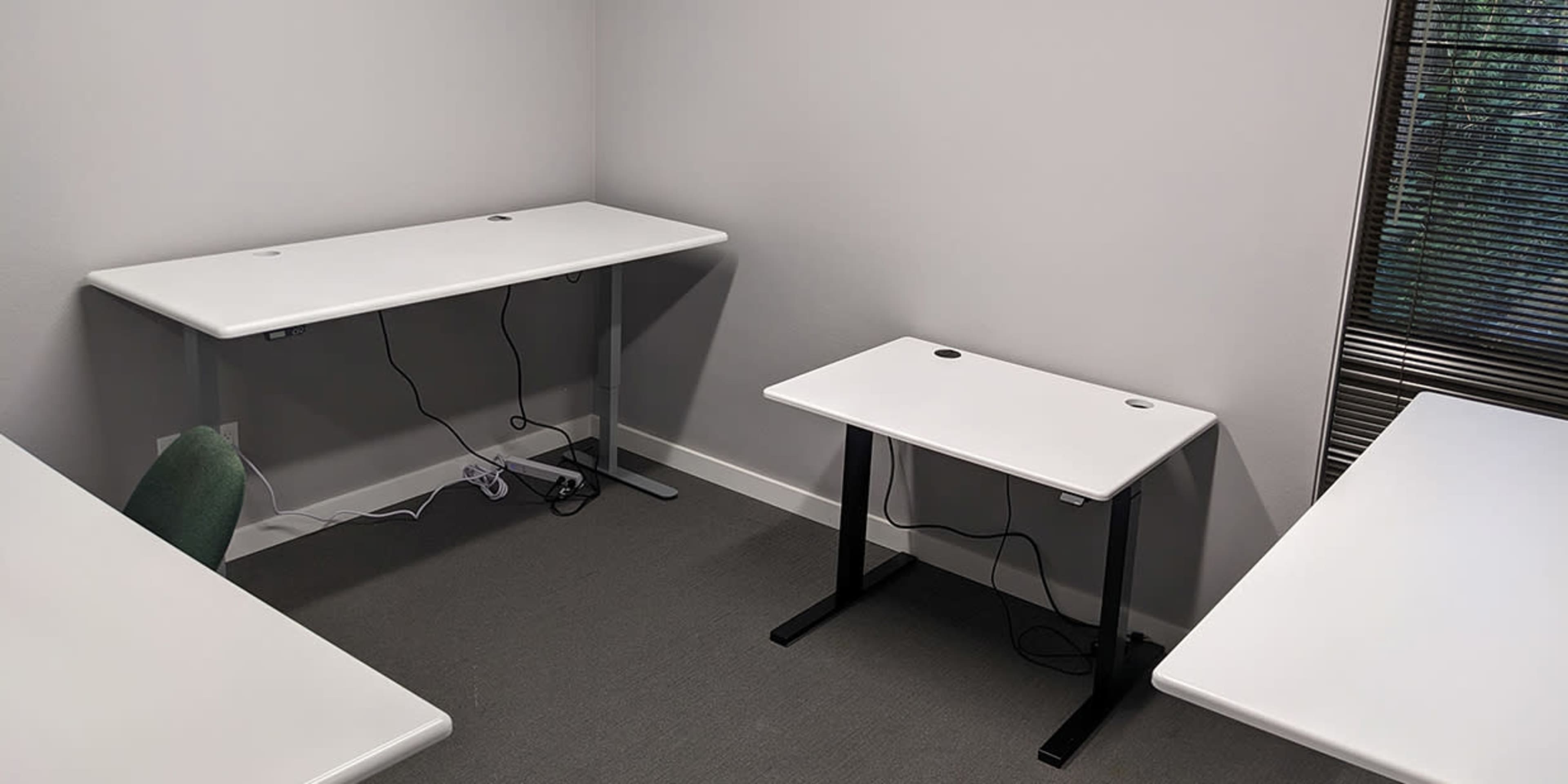 An empty room with three white desks arranged around the space, and a window with blinds on one wall.