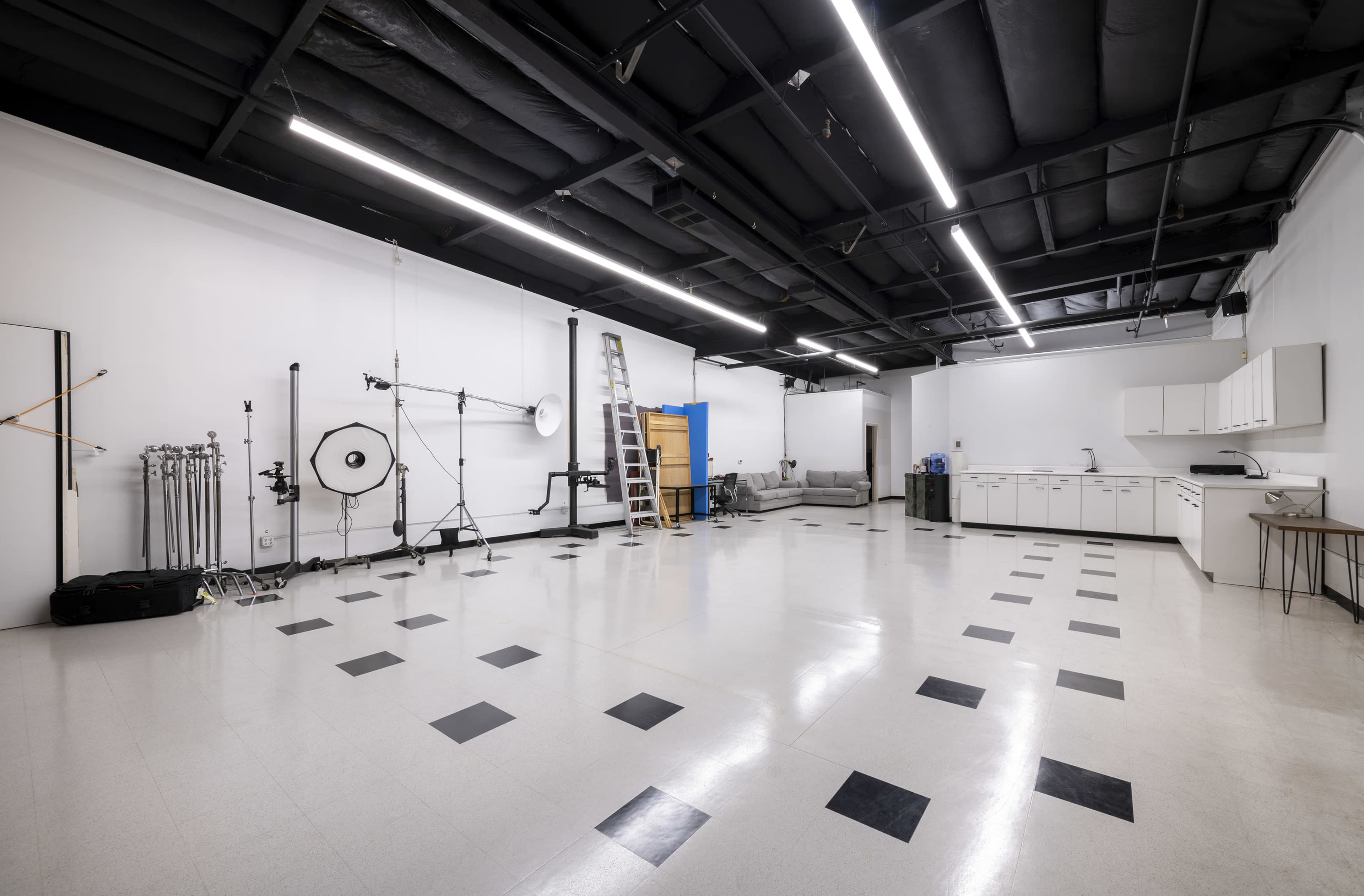The image shows a spacious studio with a polished floor, equipped with photography equipment, a ladder, and a sectional sofa against a white wall.
