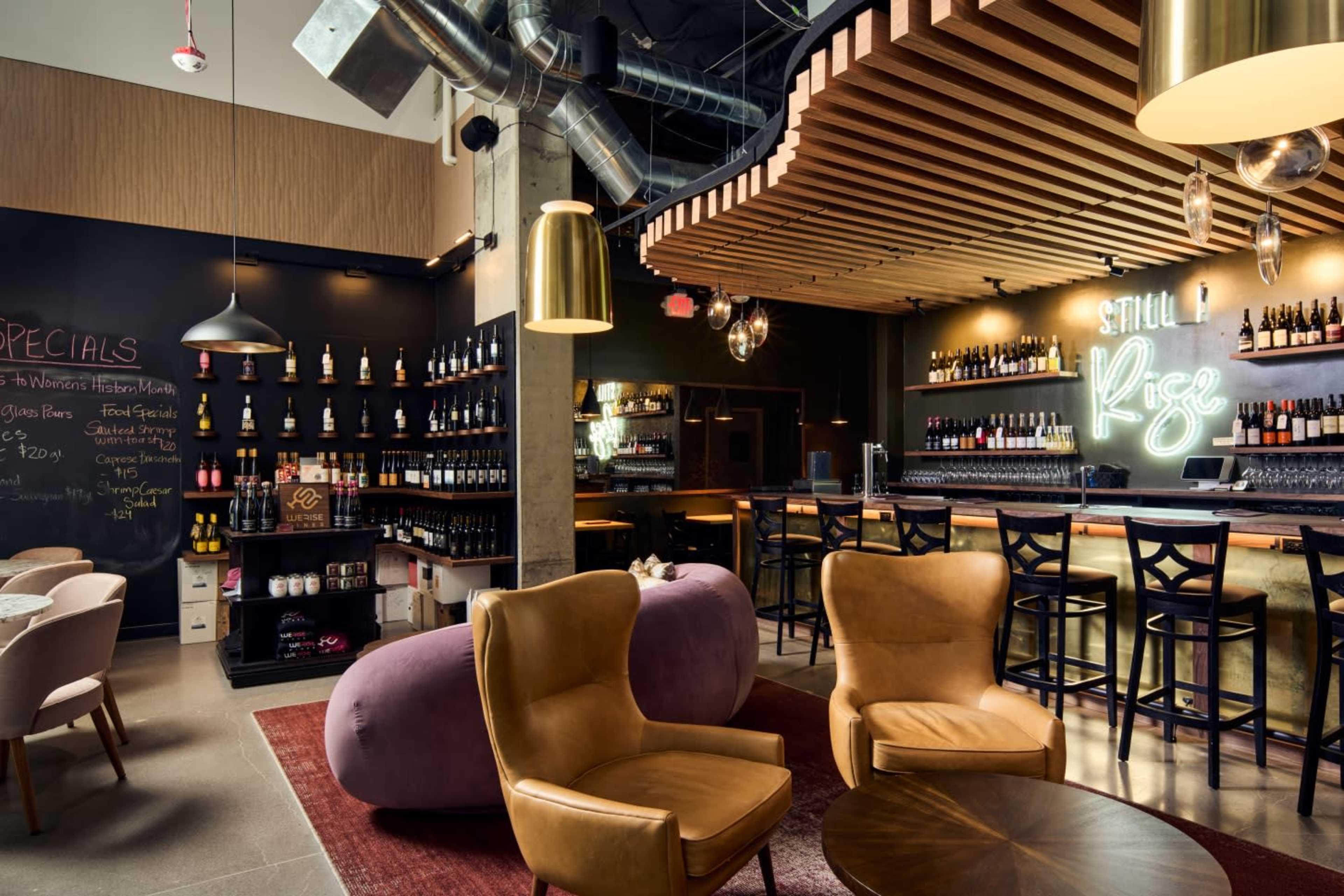 The image shows a modern bar interior with a wooden ceiling, a sleek bar area stocked with bottles, and cozy seating arrangements.