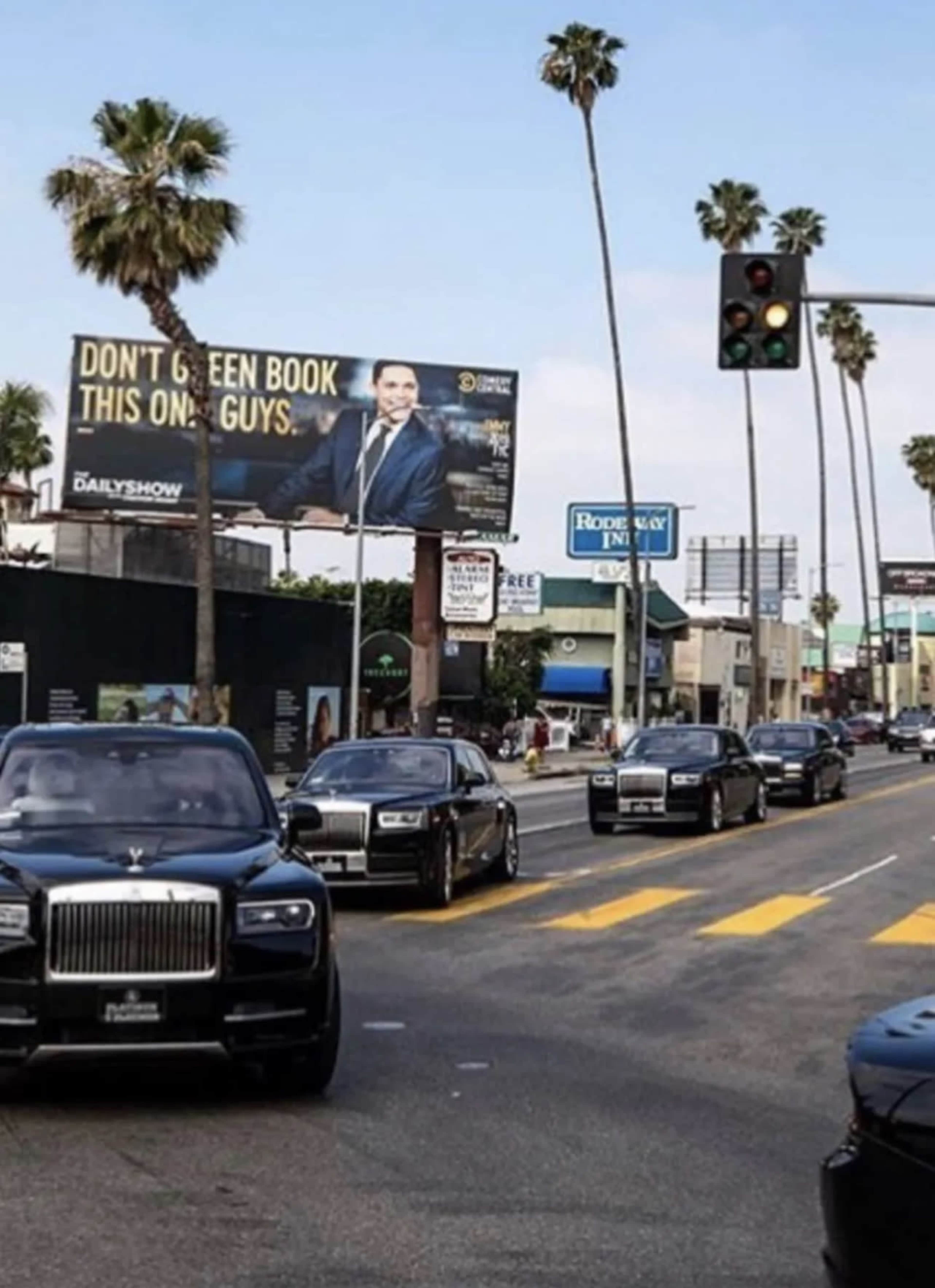 A line of black cars drives along a palm-lined street with a large billboard in the background.