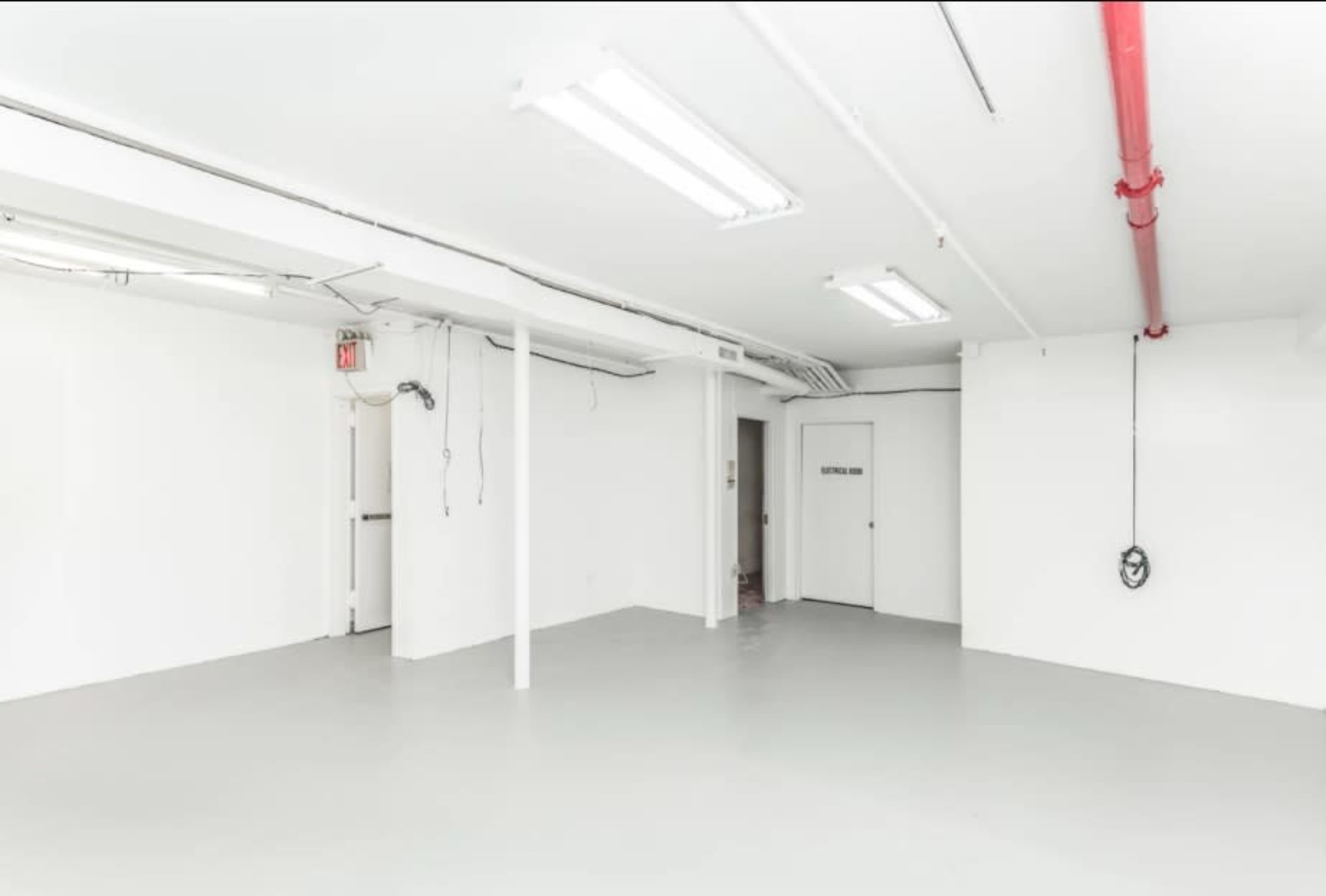 A spacious, empty room with white walls and a gray floor, featuring overhead fluorescent lights and a red pipe along the ceiling.