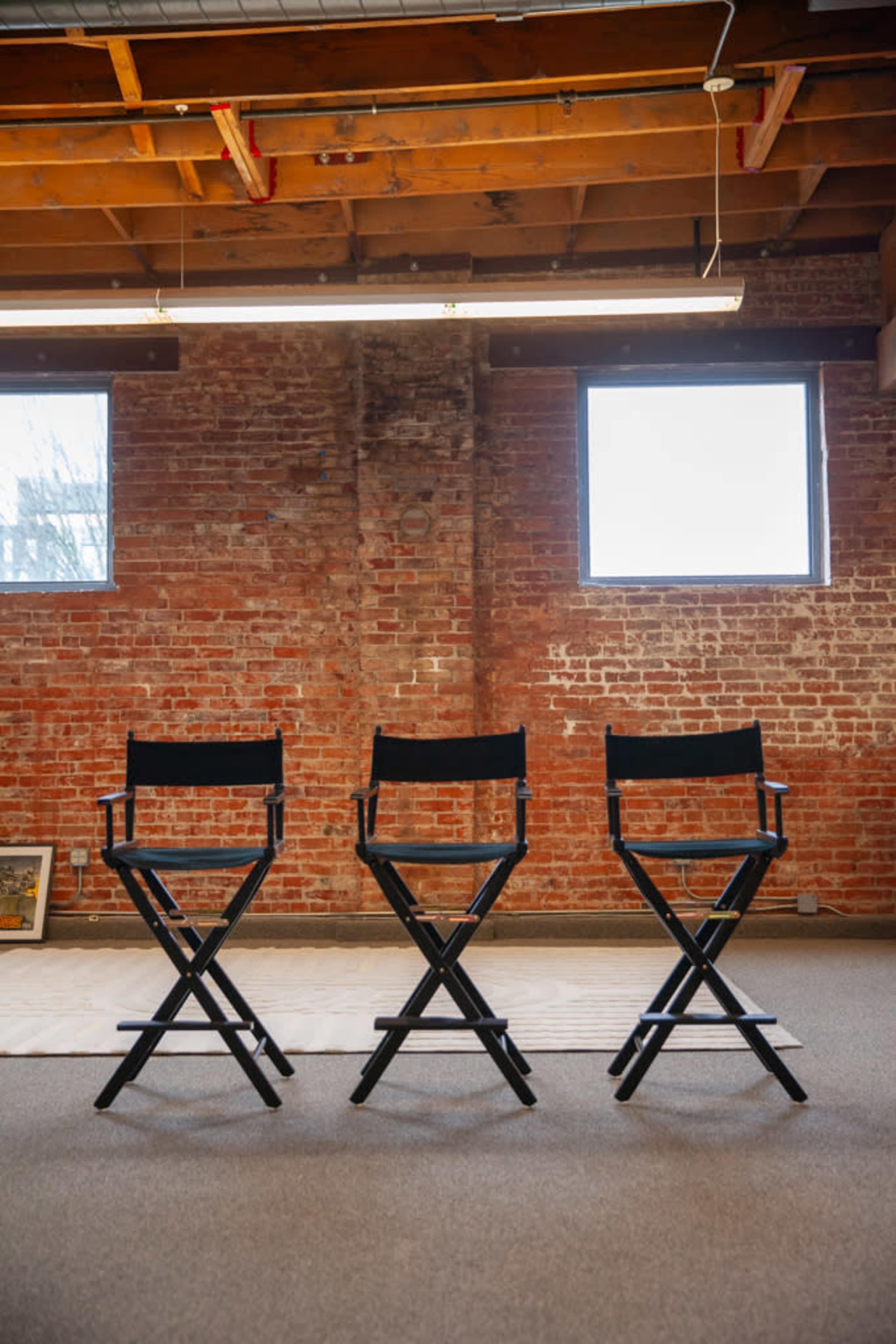 Three black director's chairs are lined up in front of large windows in a brick-walled room with a gray carpet.