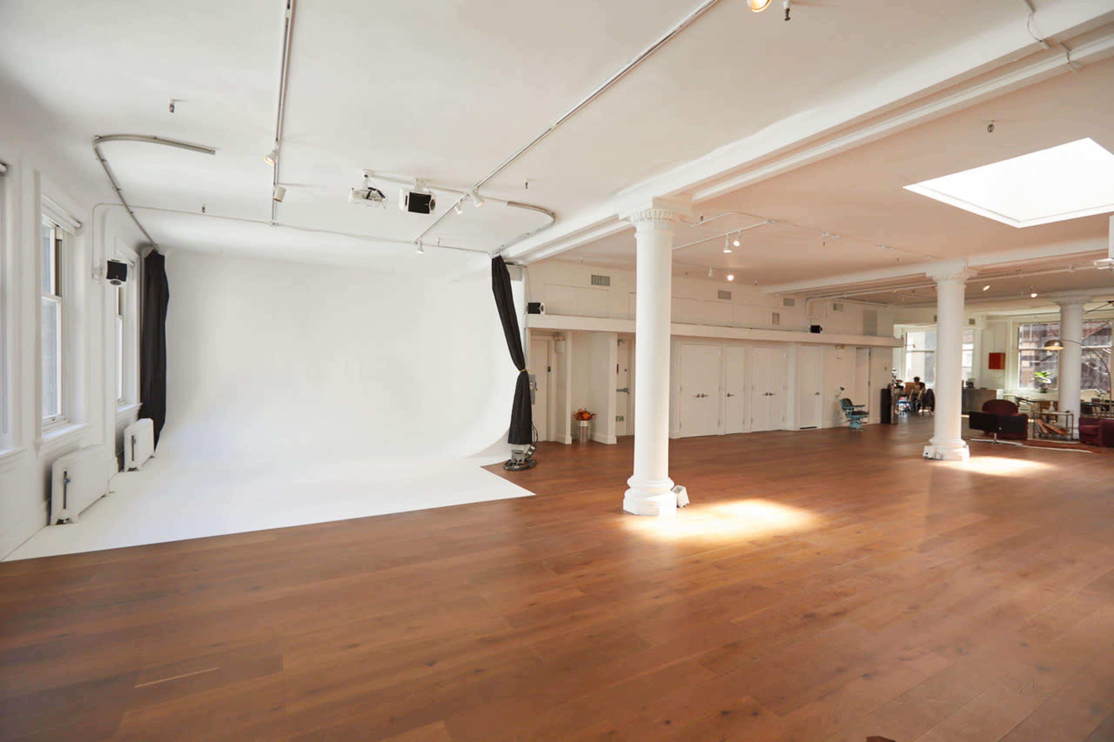 The image shows a bright, spacious studio with wooden flooring, featuring a white backdrop and several windows.