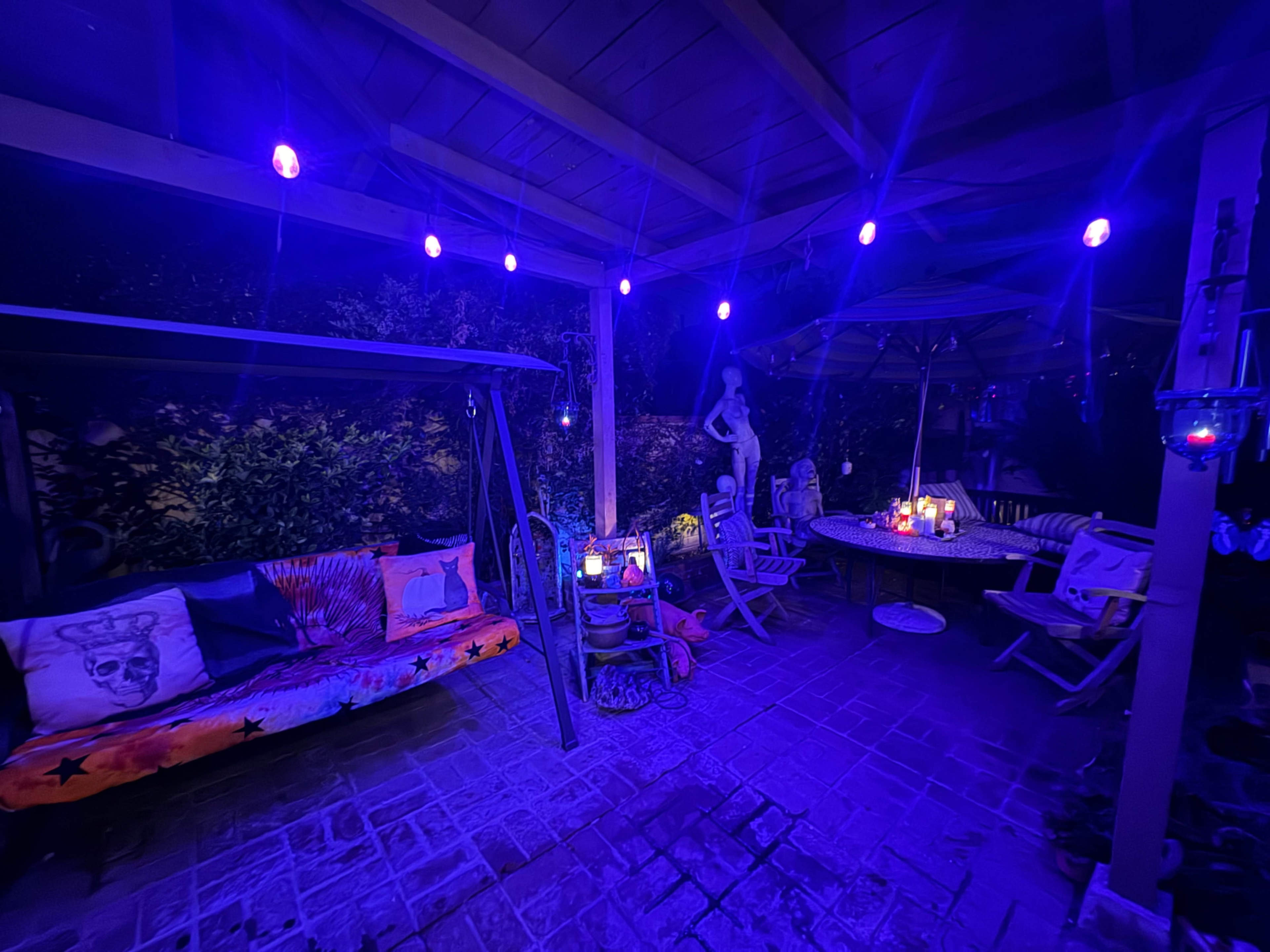 The image shows a dimly lit outdoor patio area with purple lighting, featuring a colorful couch, a dining table, and decorative elements.