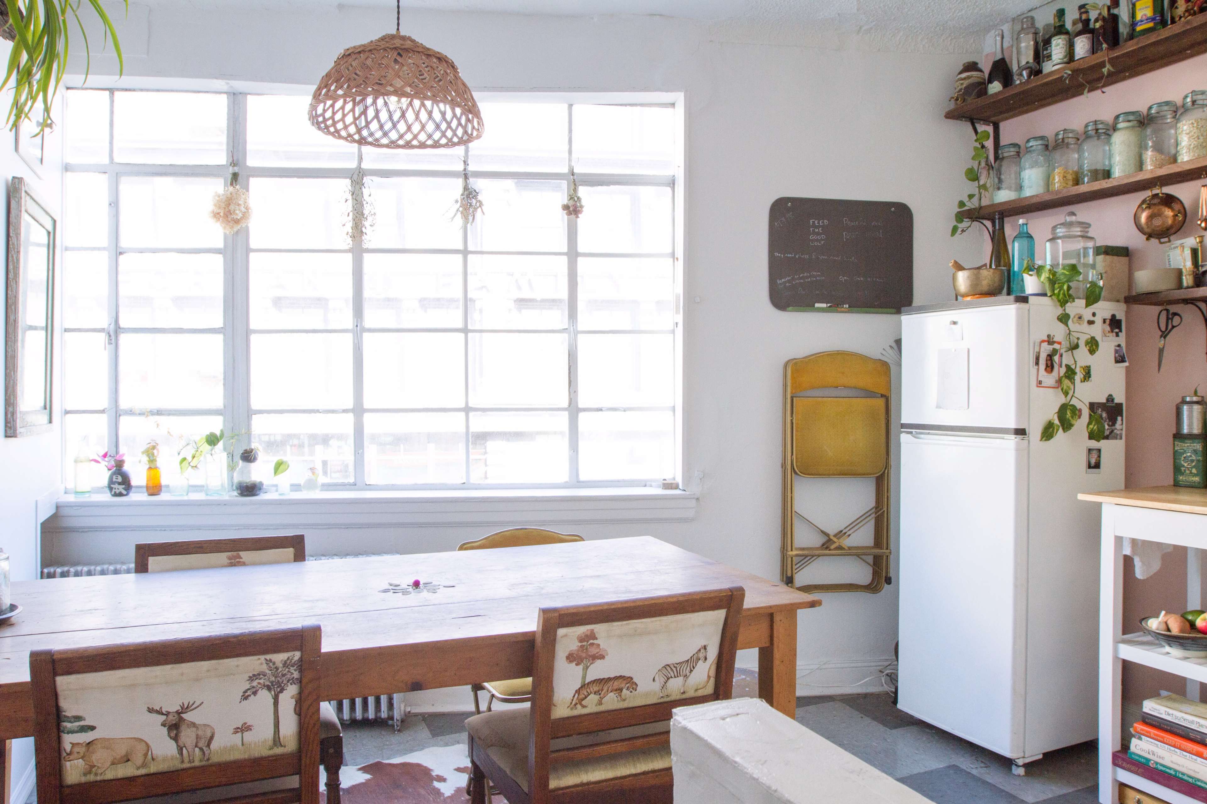 A bright kitchen with a wooden dining table, metal chairs, a white refrigerator, and large windows allowing natural light to enter the space.