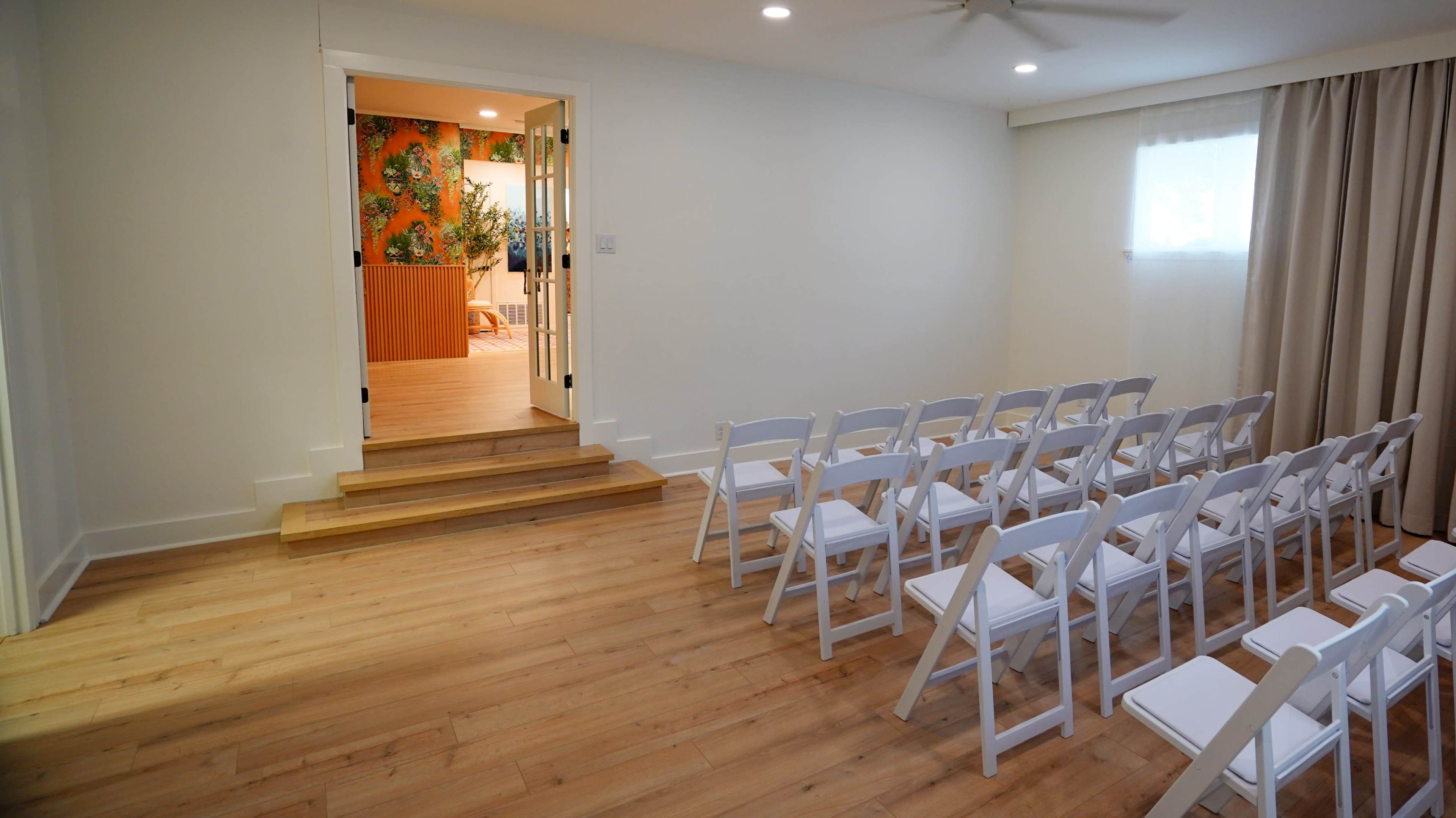 The scene features a small, bright room with white walls and wooden flooring, furnished with rows of white folding chairs facing an open doorway leading to a decorated area with vibrant wallpaper.
