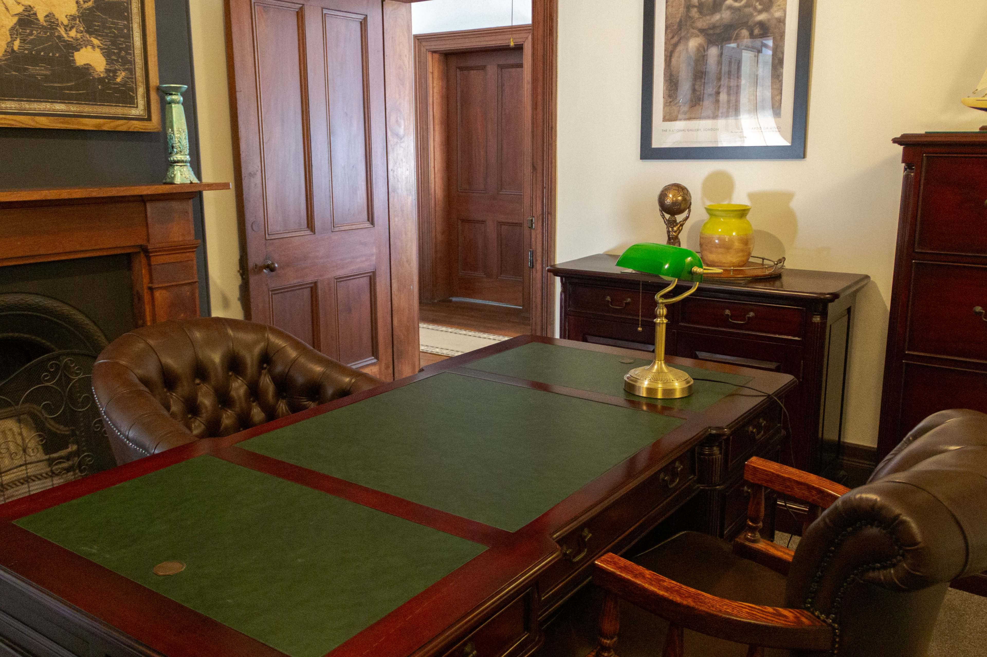 The image shows a vintage wooden office with a large desk featuring a green leather top, a brass lamp, and two leather chairs, along with a fireplace and doorways leading to other rooms.