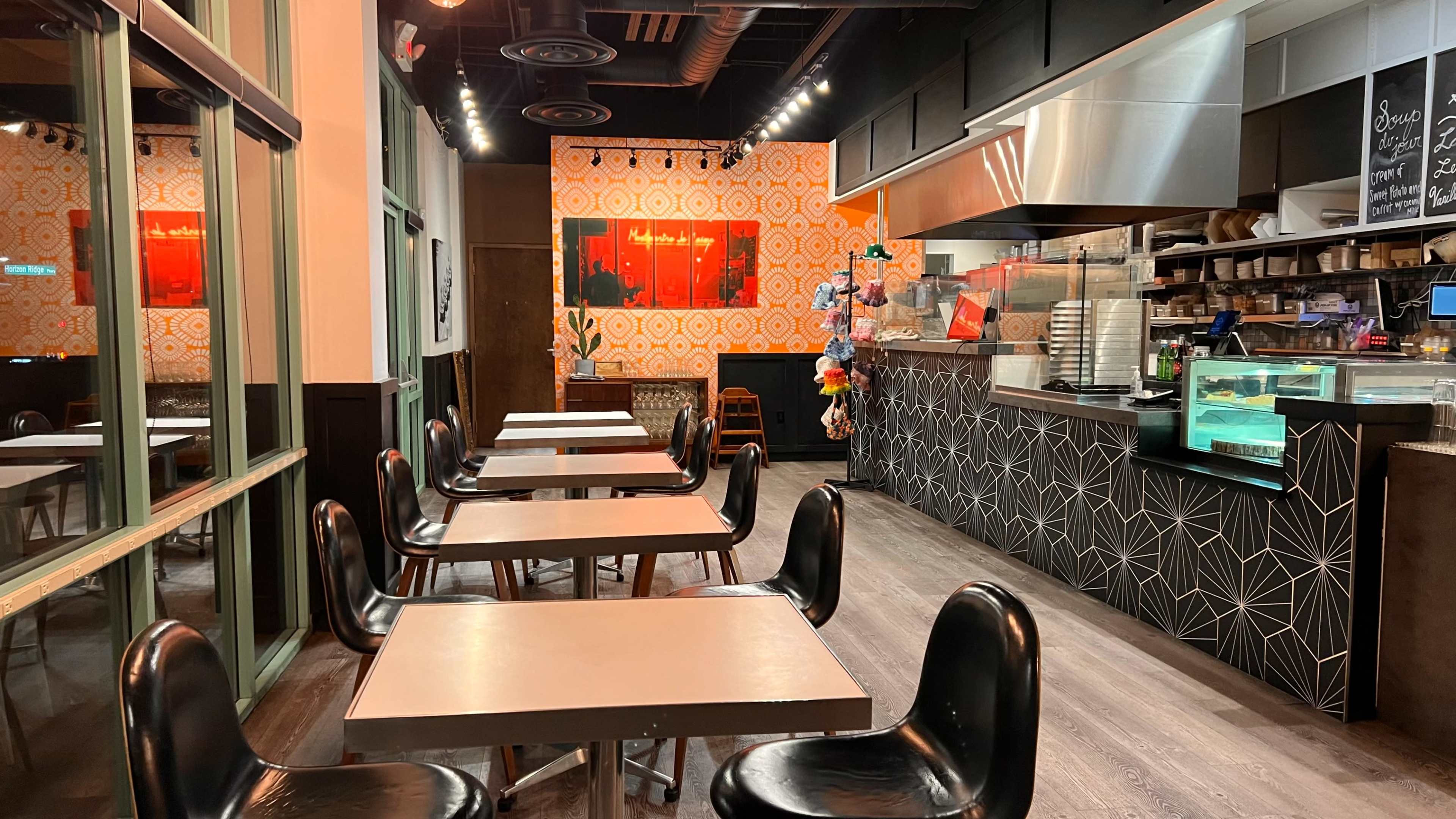An empty restaurant interior with black chairs and tables, a patterned orange wall, and a service counter displaying food items.