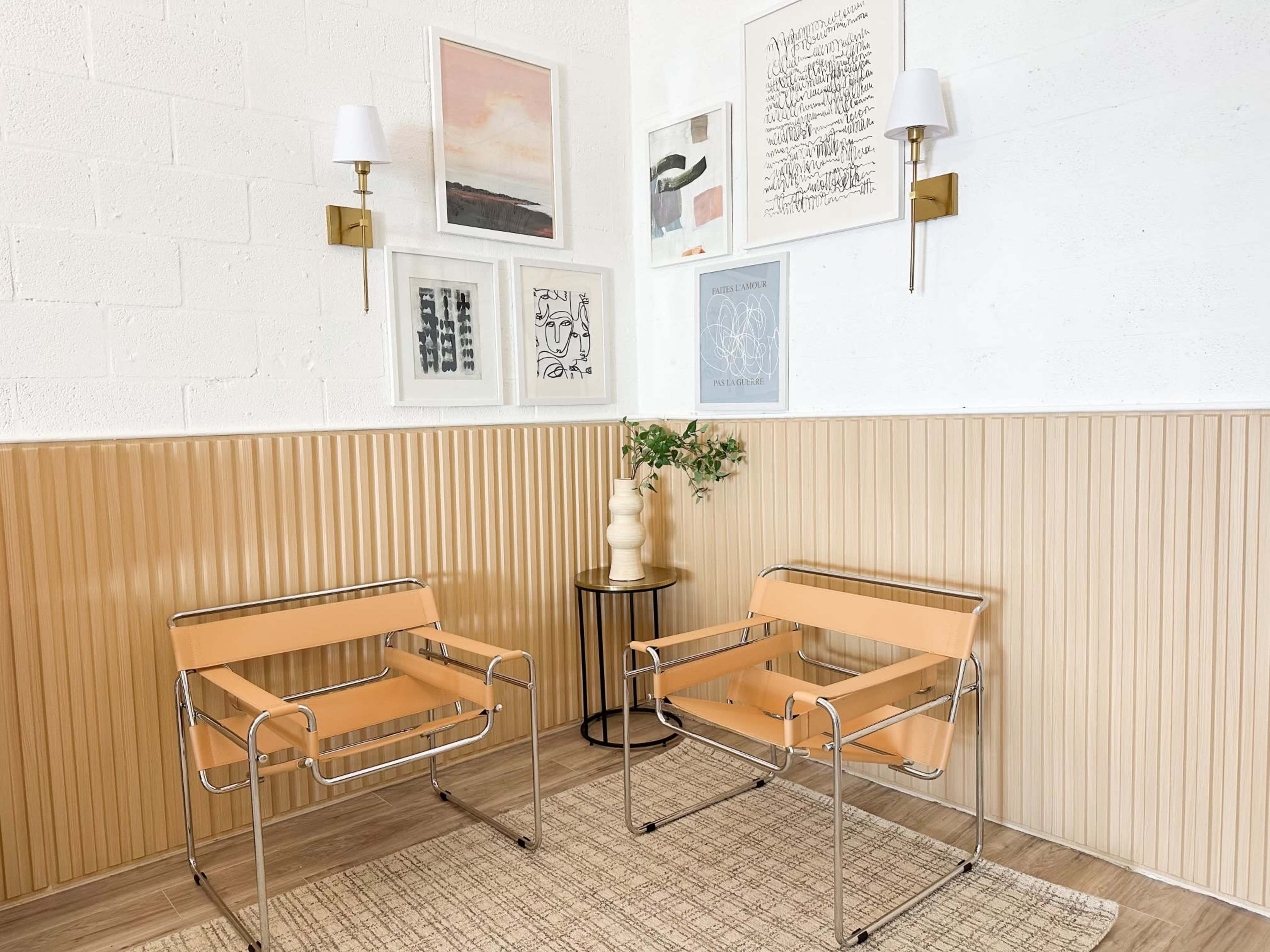 Two modern chairs with tan cushions are positioned on a textured rug beside a small table, against a backdrop of framed artwork and a paneled wall.