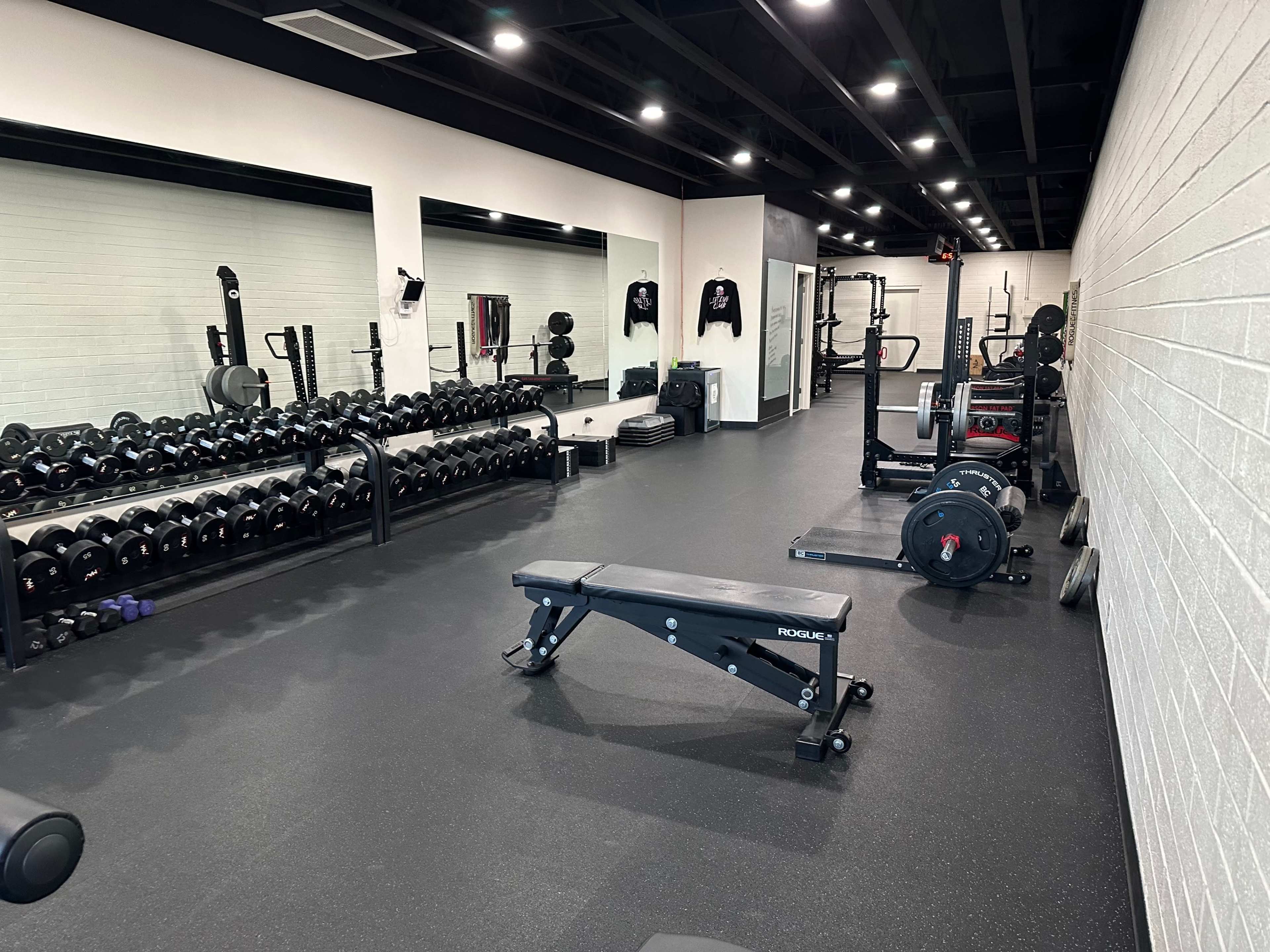 The image shows a well-equipped gym with a row of dumbbells, a weight bench, and various strength training equipment in a spacious layout.