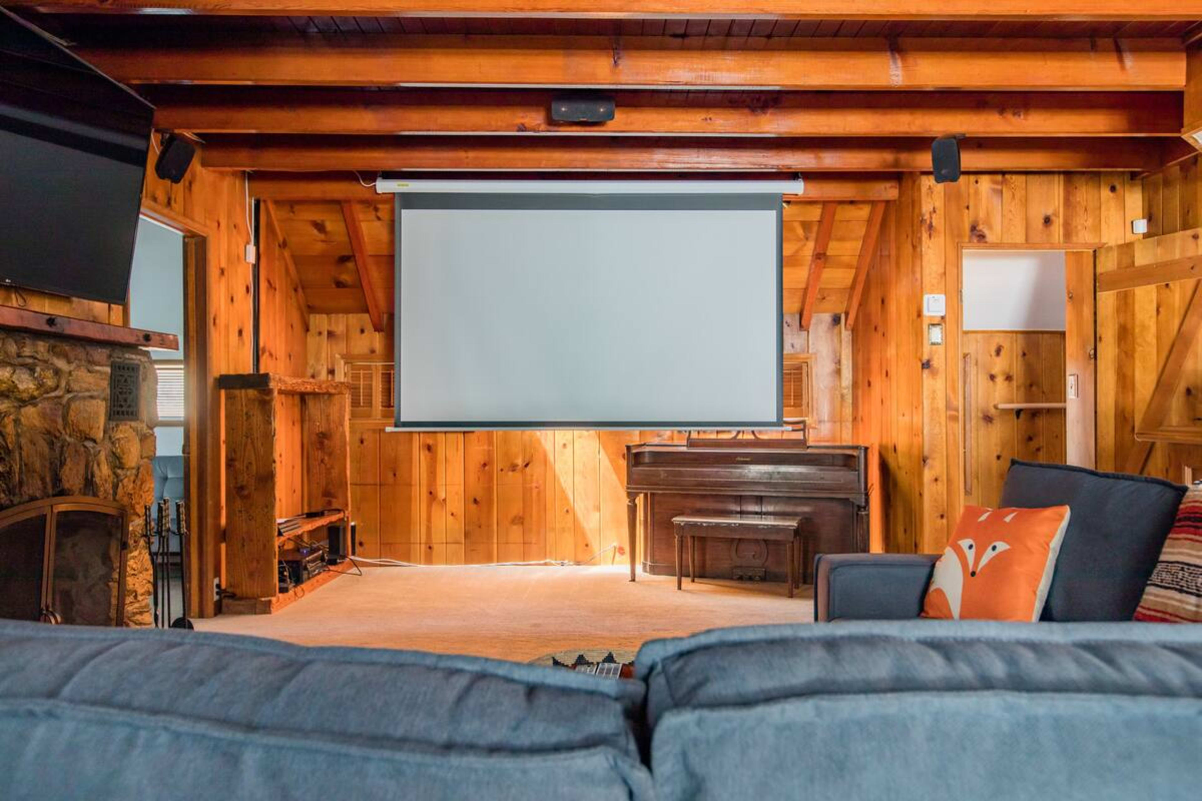 The image shows a rustic living room with wooden walls, a large projection screen, a piano, and comfortable seating.