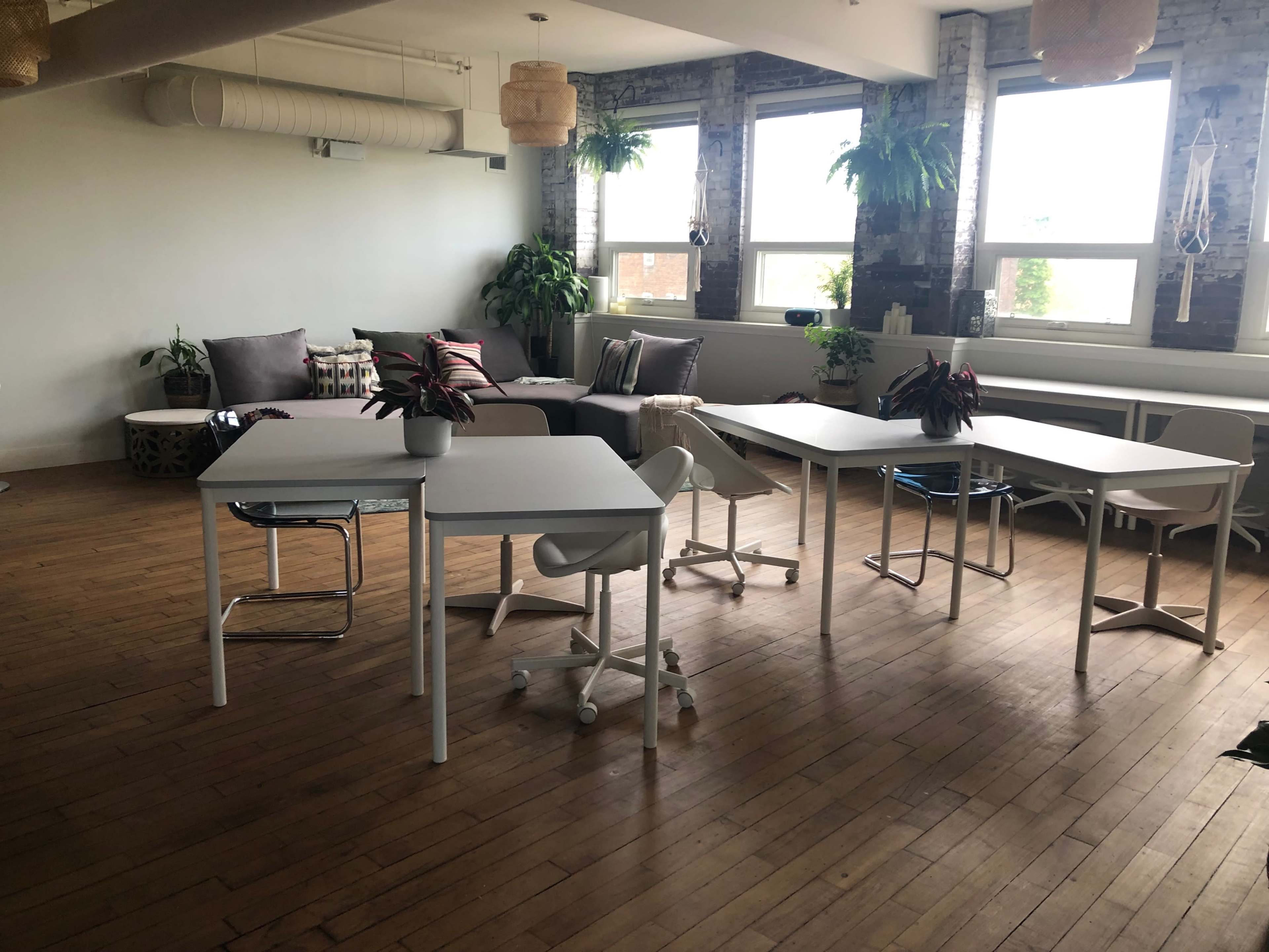 The image shows a spacious room with wooden flooring, featuring several white desks and chairs arranged around plants and a seating area.