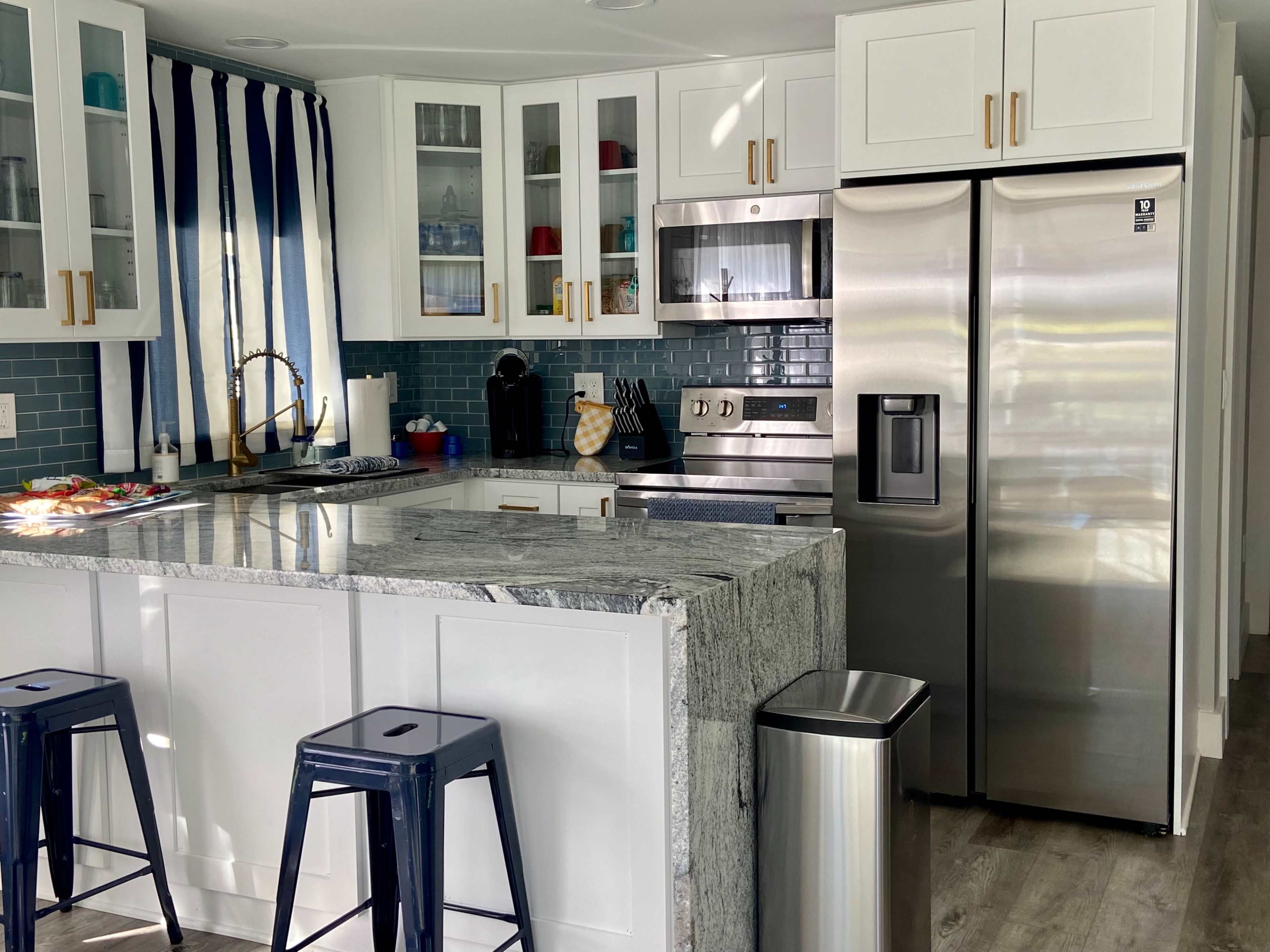 A modern kitchen with a white island, stainless steel appliances, blue and white cabinetry, and a bar area with two metal stools.