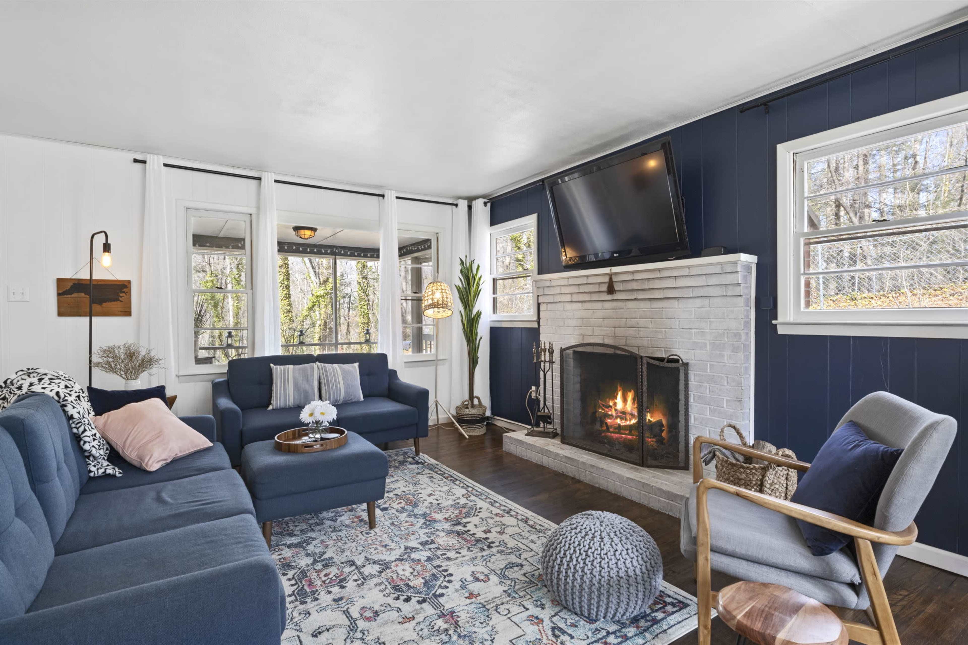 The living room features a fireplace with a TV above it, flanked by two windows, and furnished with a blue sofa set, a patterned rug, and a cozy armchair.