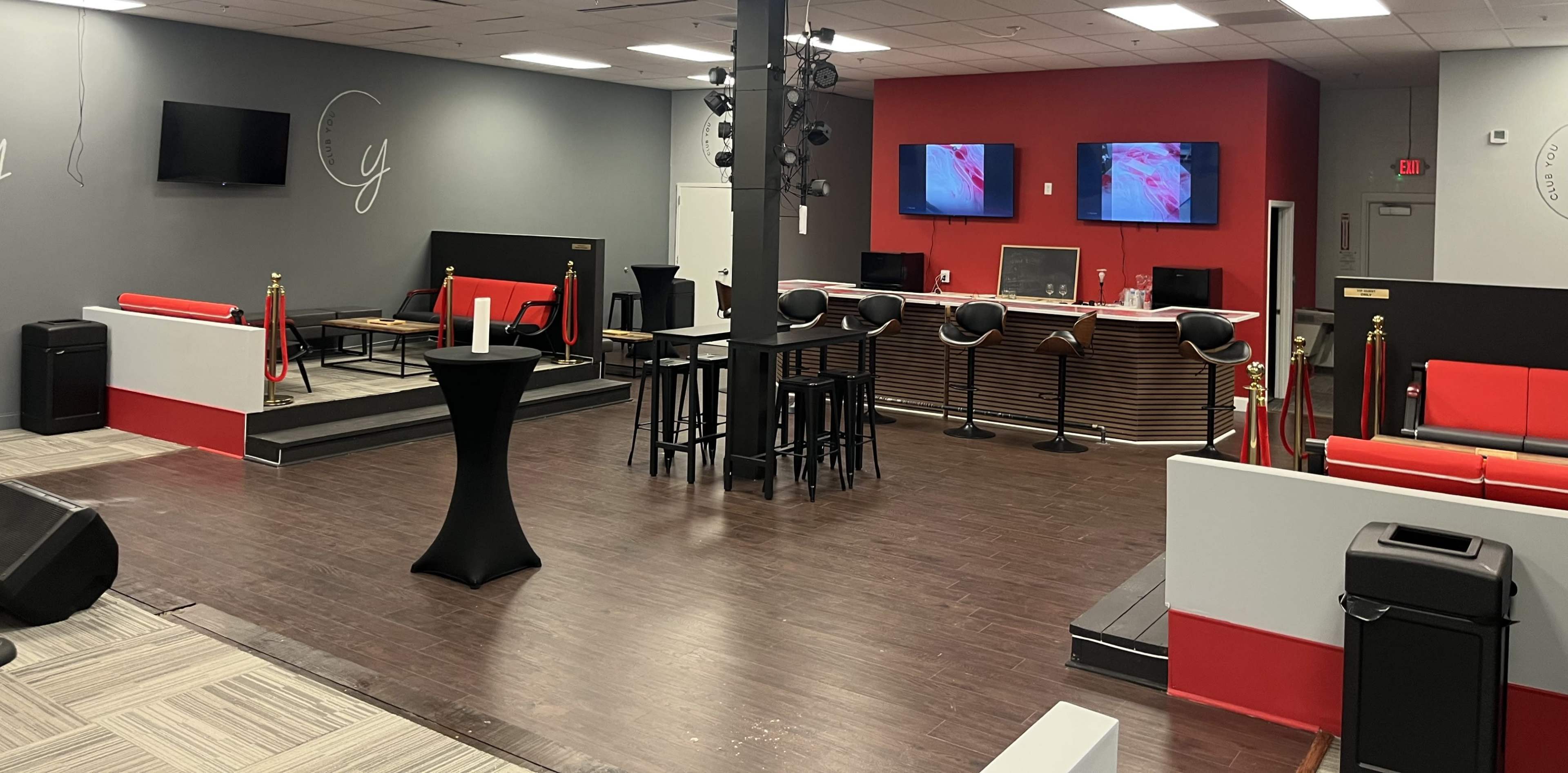 The image shows a spacious interior of a venue featuring seating areas, a bar counter, and multiple screens on the walls.