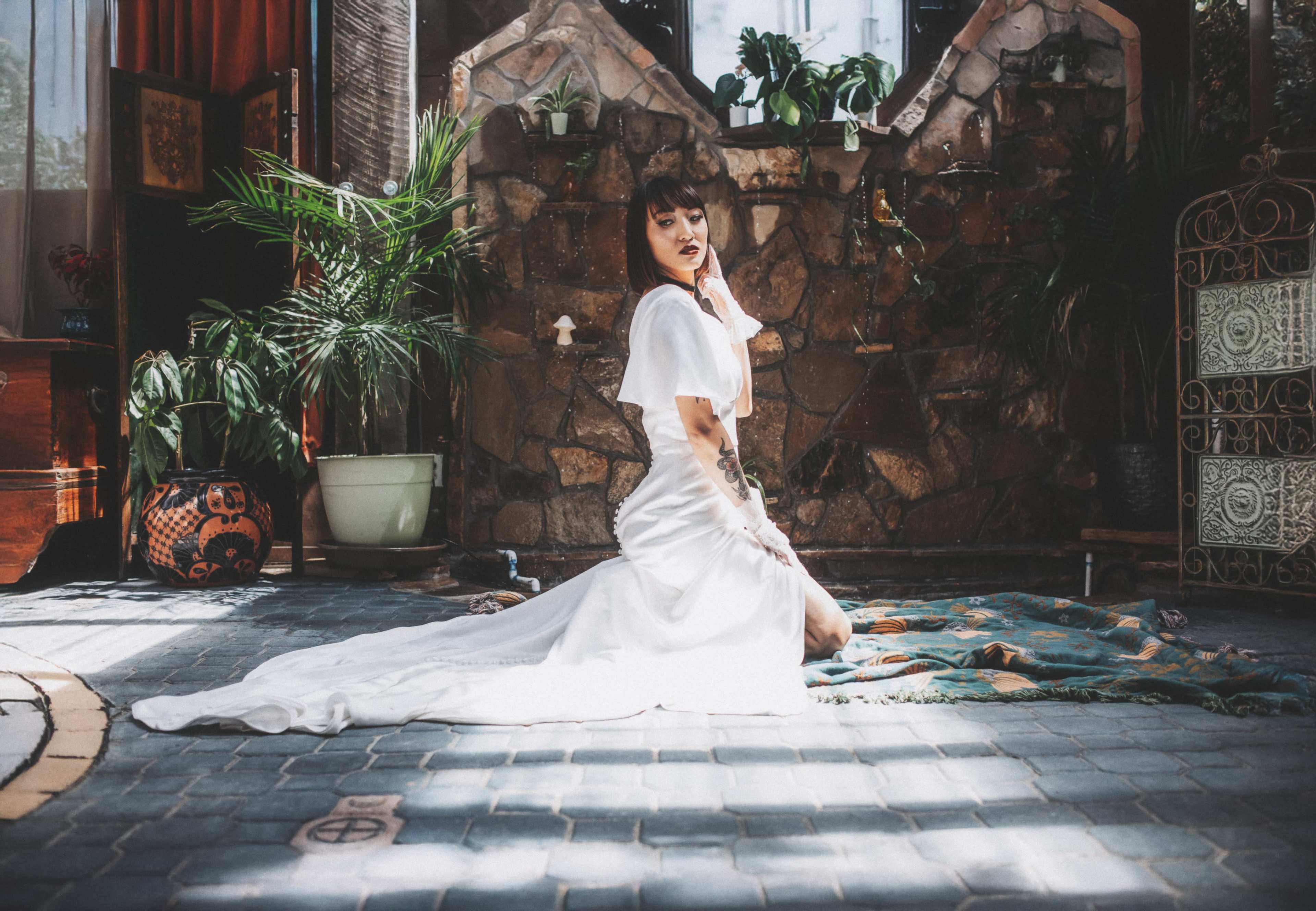 A woman in a white dress poses on a patterned mat in a sunlit room adorned with plants and stone walls.