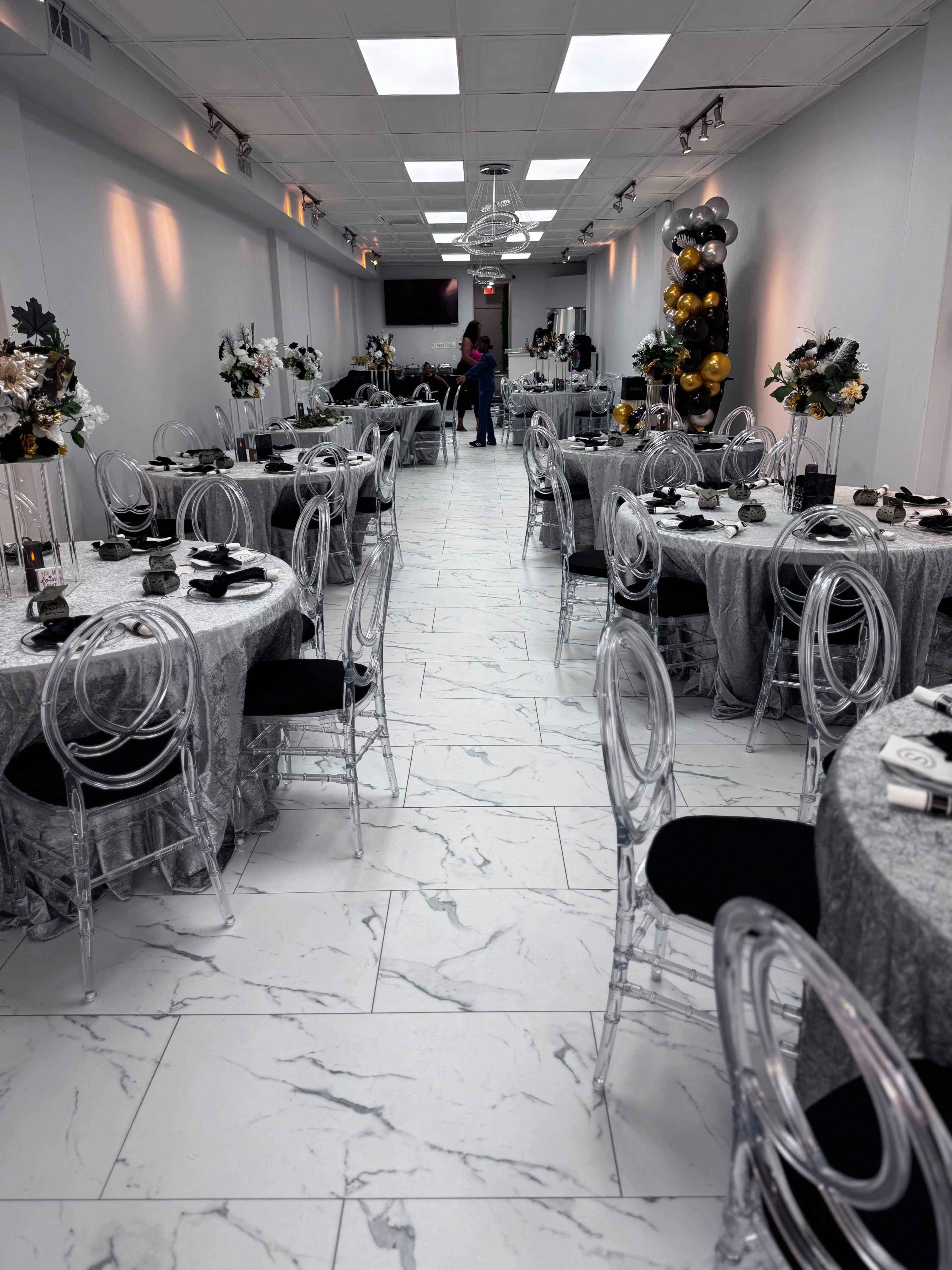 The image shows a modern event space with decorated round tables covered in silver tablecloths and clear chairs, arranged neatly along a marble-tiled floor.