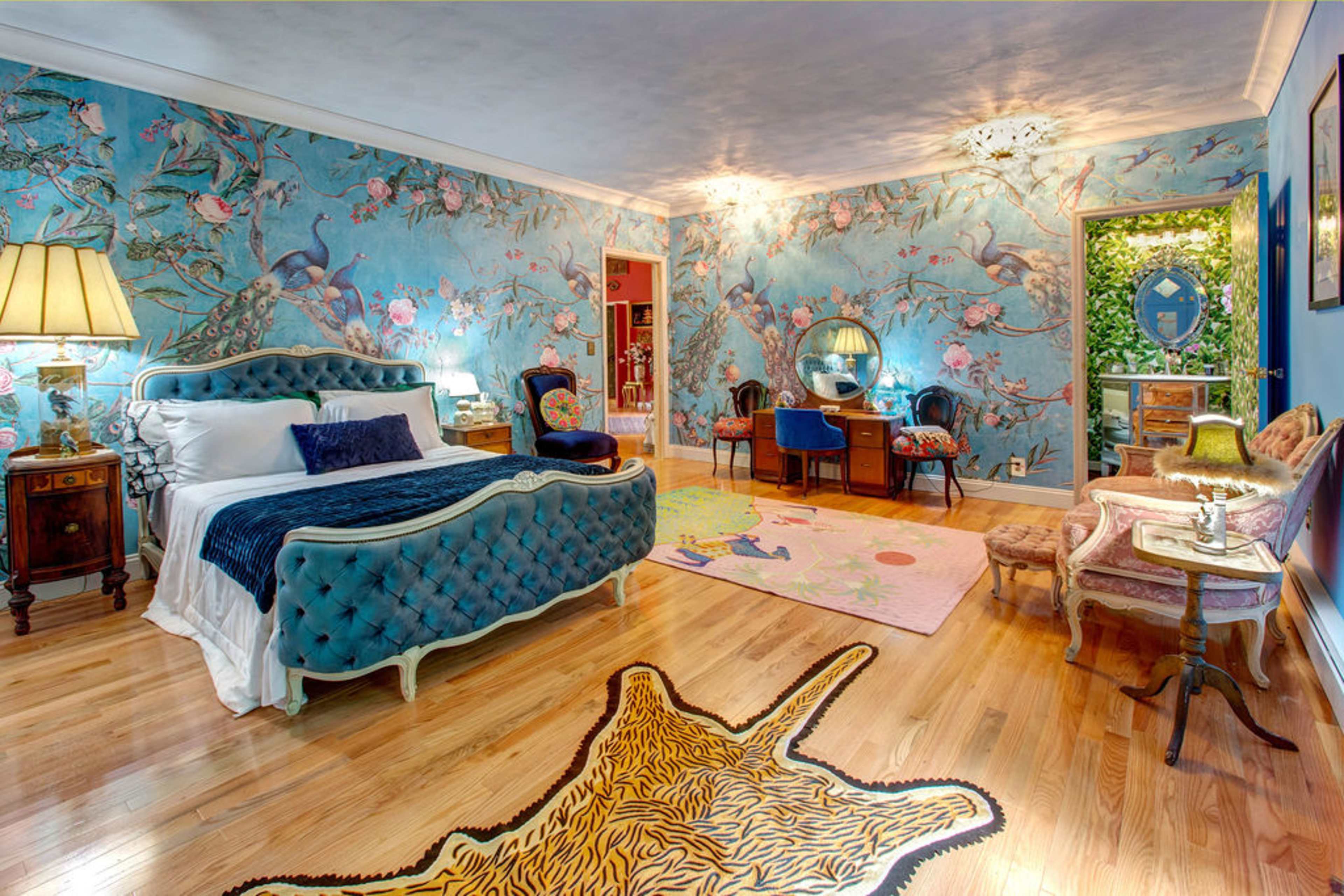 The image shows a brightly decorated bedroom featuring a blue floral wall mural, a tufted bed, an ornate dresser, and a tiger-striped rug on the wooden floor.