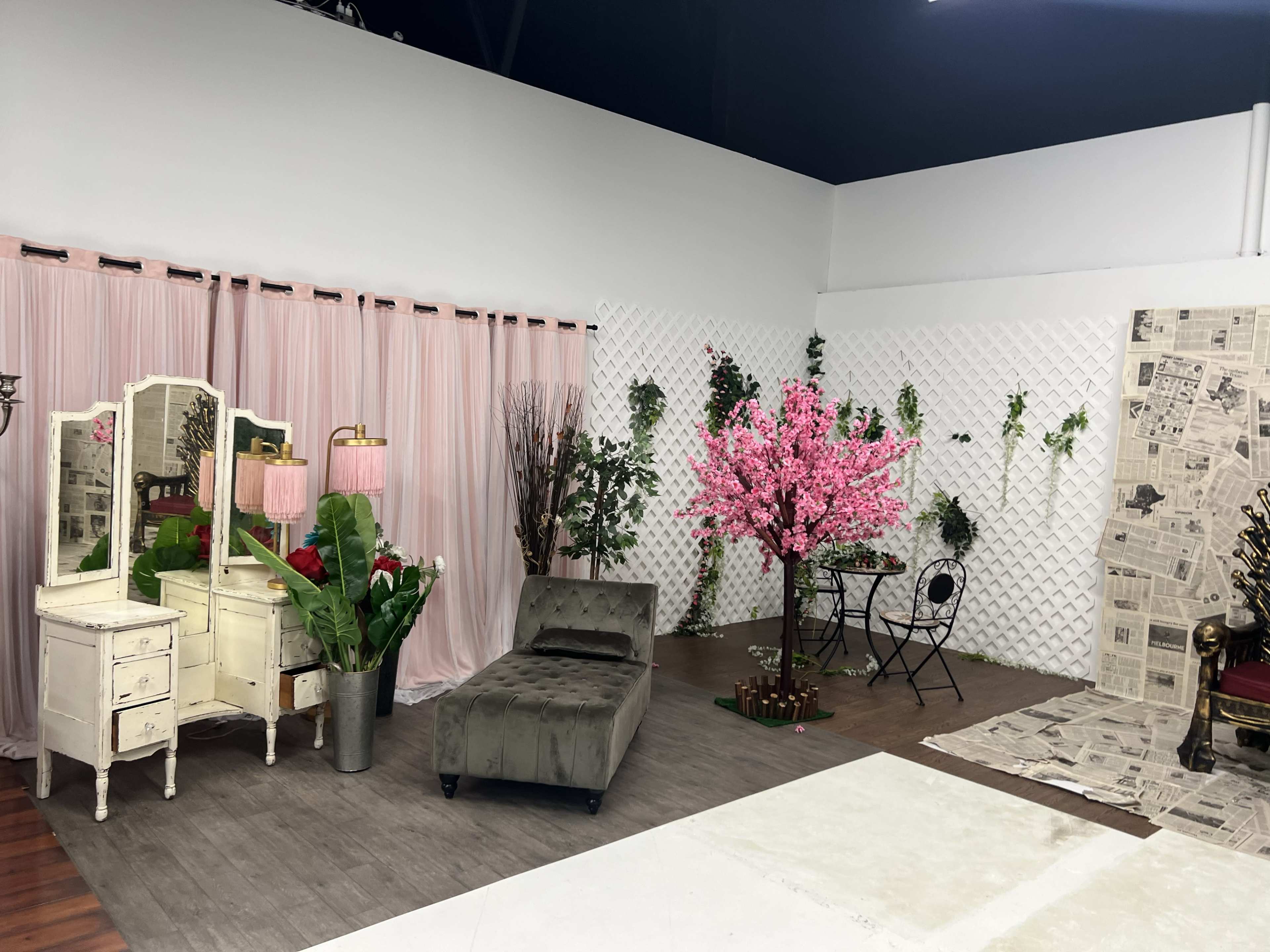 The image shows a staged room featuring a vintage dresser, a gray chaise lounge, a small table with chairs, and a cherry blossom tree among decorative elements and light pink curtains.