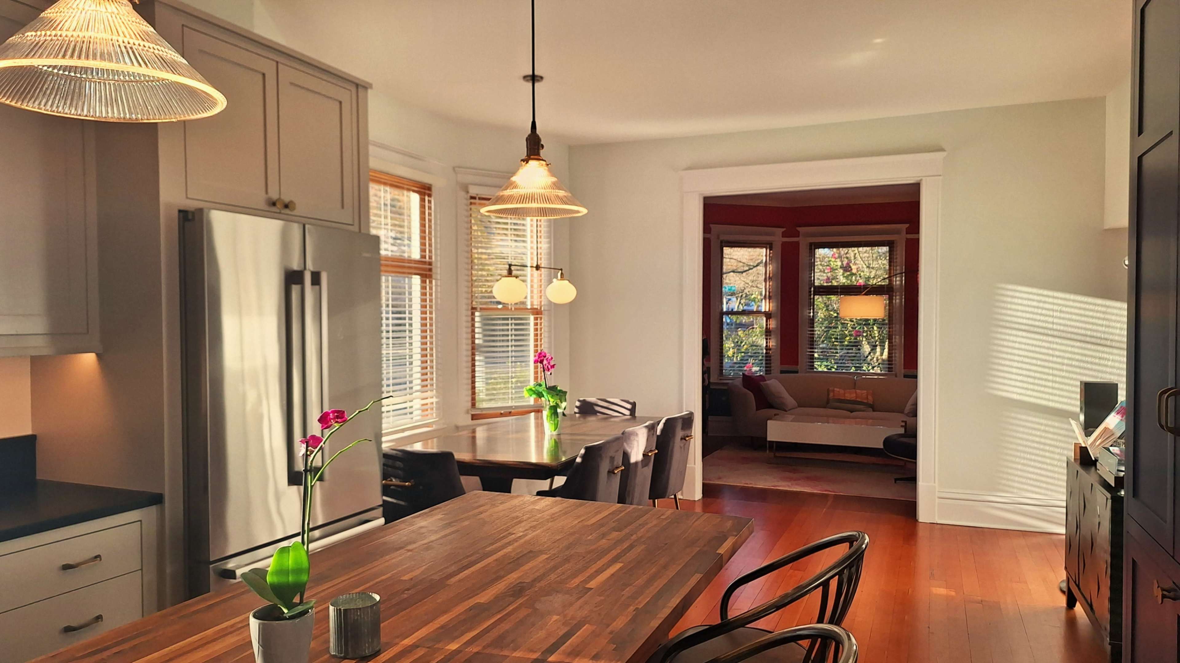 The image shows a bright kitchen with a wooden dining table, modern appliances, and large windows letting in natural light, leading into a cozy living area with a couch.