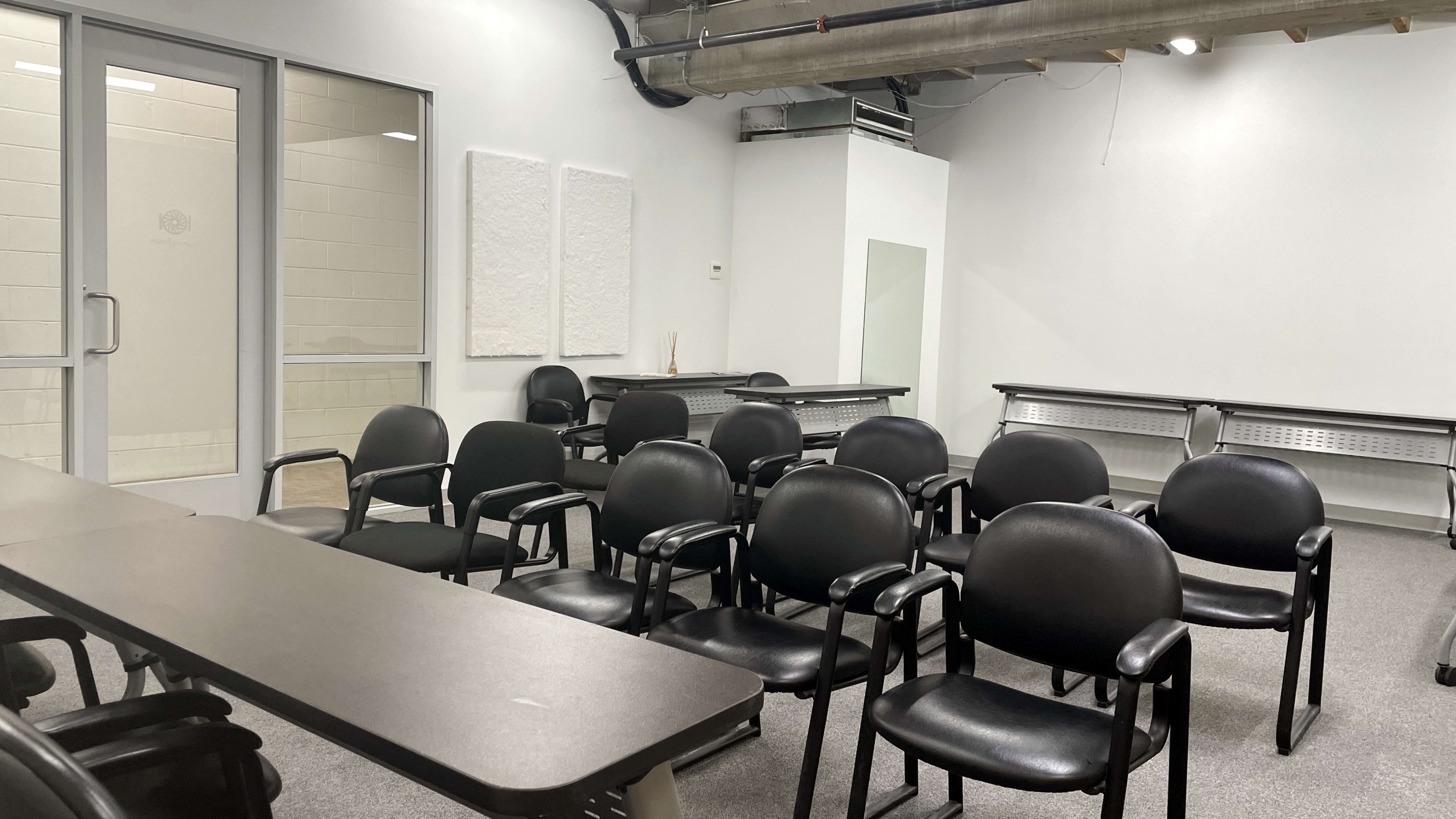 A meeting room with several black chairs arranged in rows, a few tables, and a partially visible glass door leading to another space.