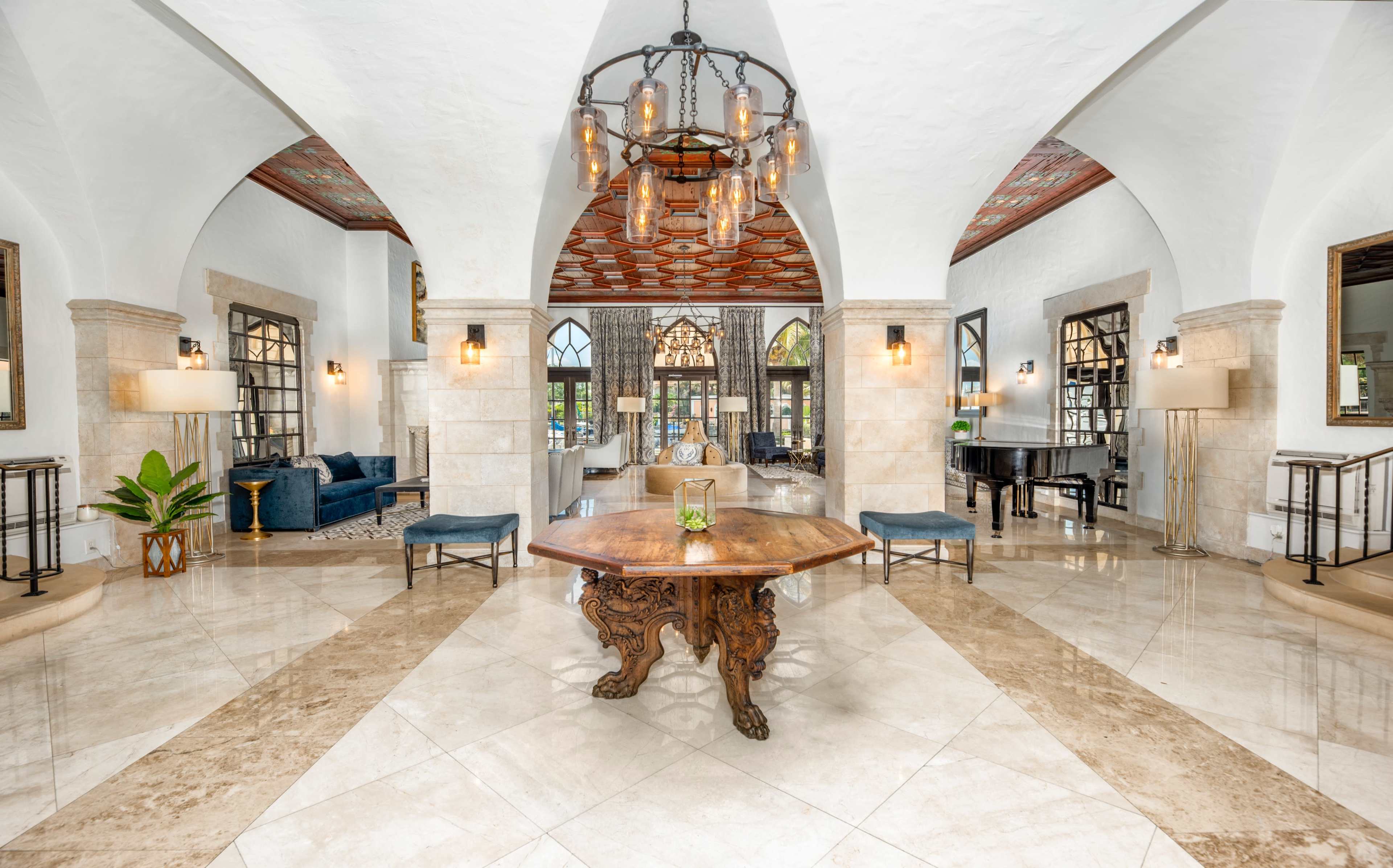 A spacious, elegantly designed lobby featuring high arched ceilings, a large wooden table at the center, and a mix of comfortable seating areas.