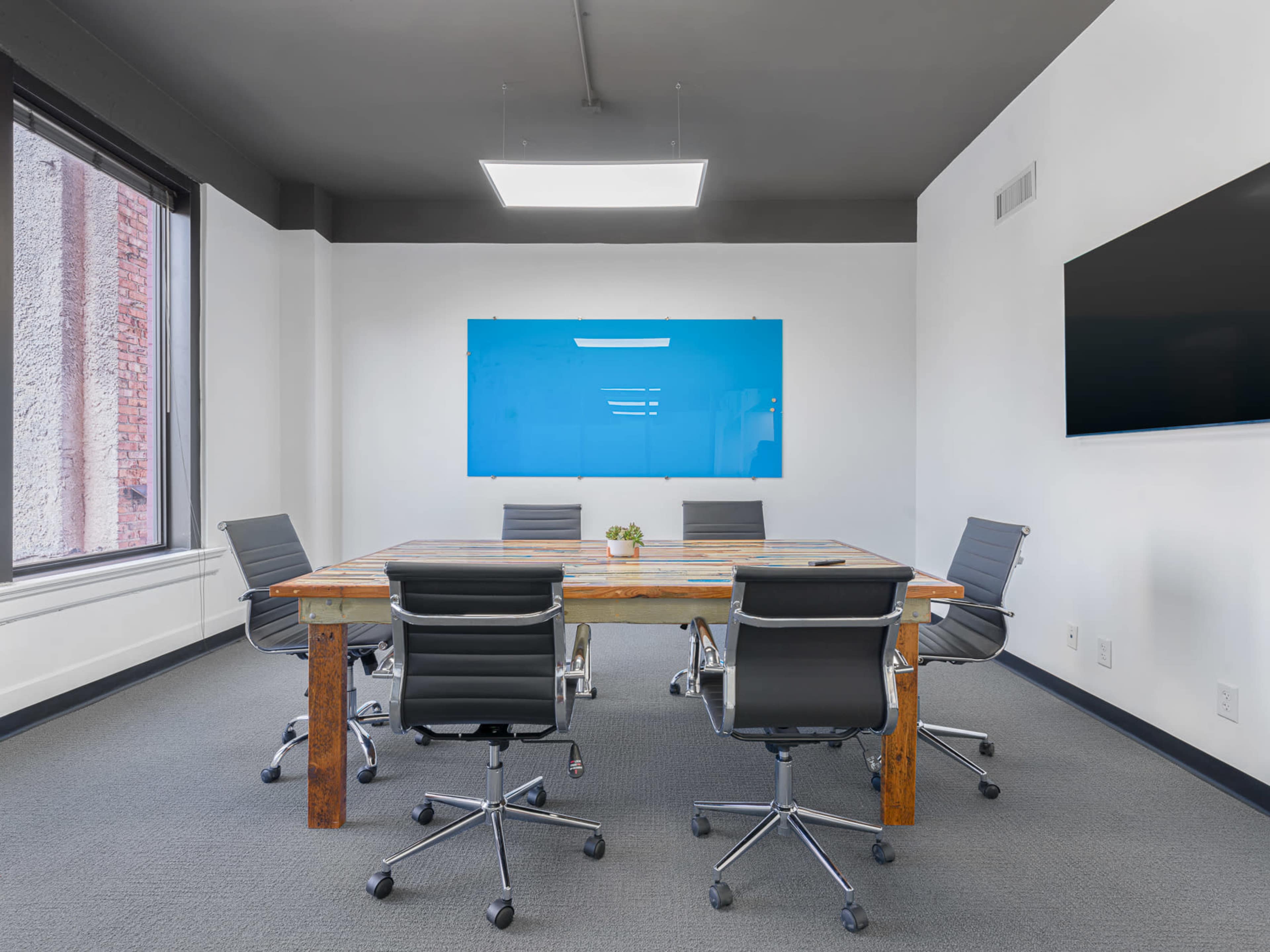 A modern conference room features a large rectangular table surrounded by five ergonomic chairs, with a large blue presentation board and a wall-mounted screen.