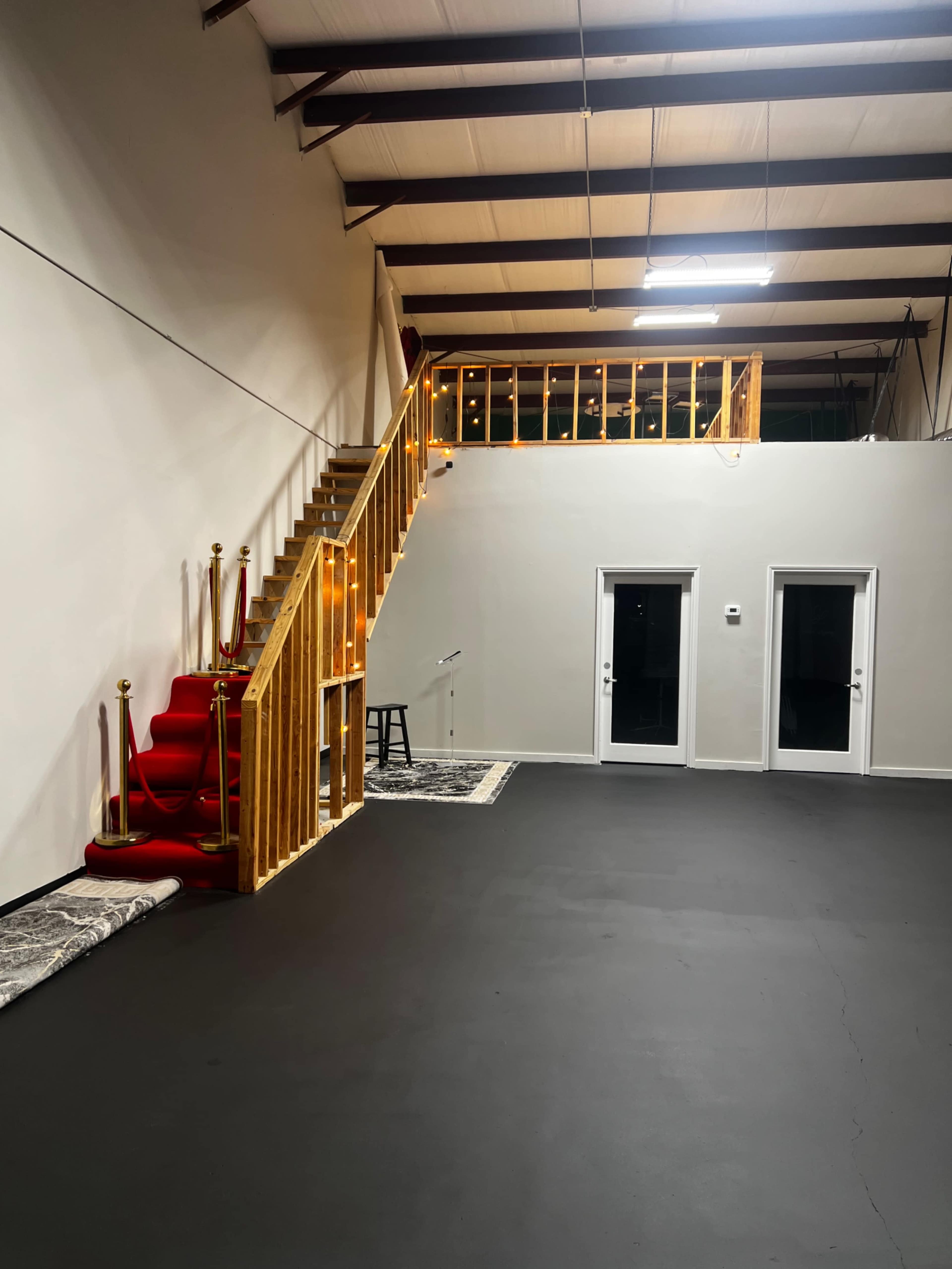 The image shows a spacious, empty interior with a red carpeted staircase leading to a loft area, along with two glass doors on the right wall.