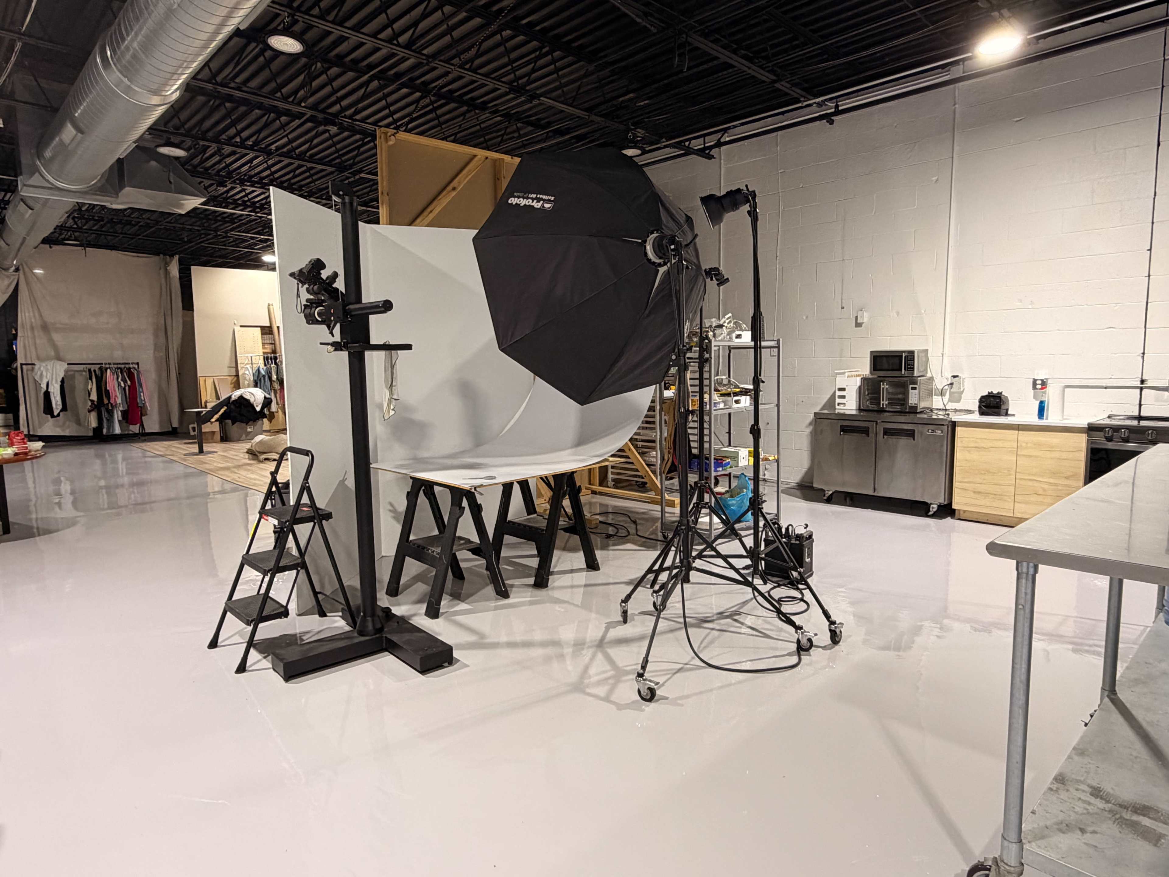 The image shows a photography studio setup with a large softbox light, a backdrop, and various equipment including a step ladder and kitchen appliances in the background.