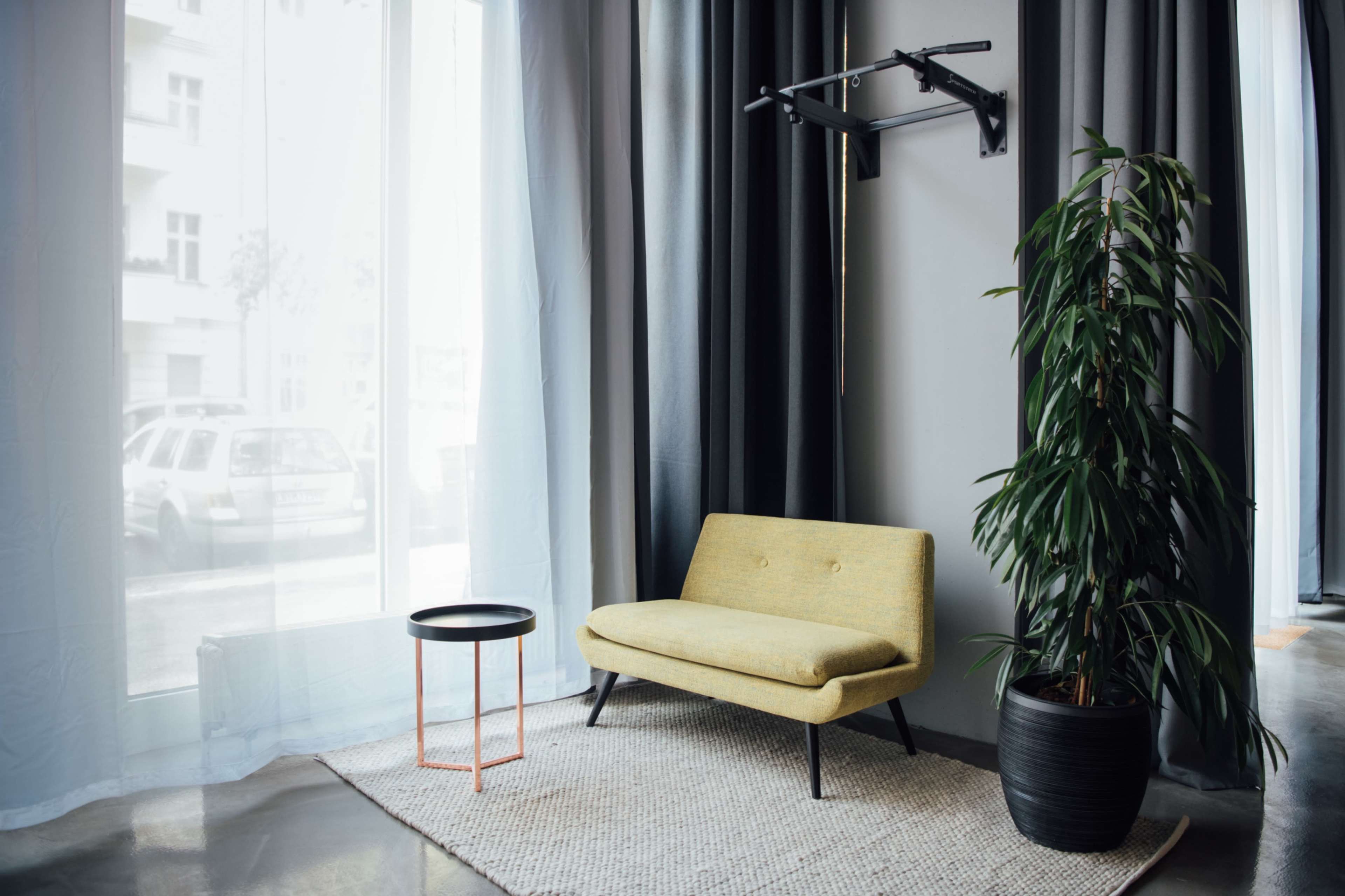 A yellow loveseat sits beside a round black table and a large potted plant in a brightly lit room with sheer curtains.