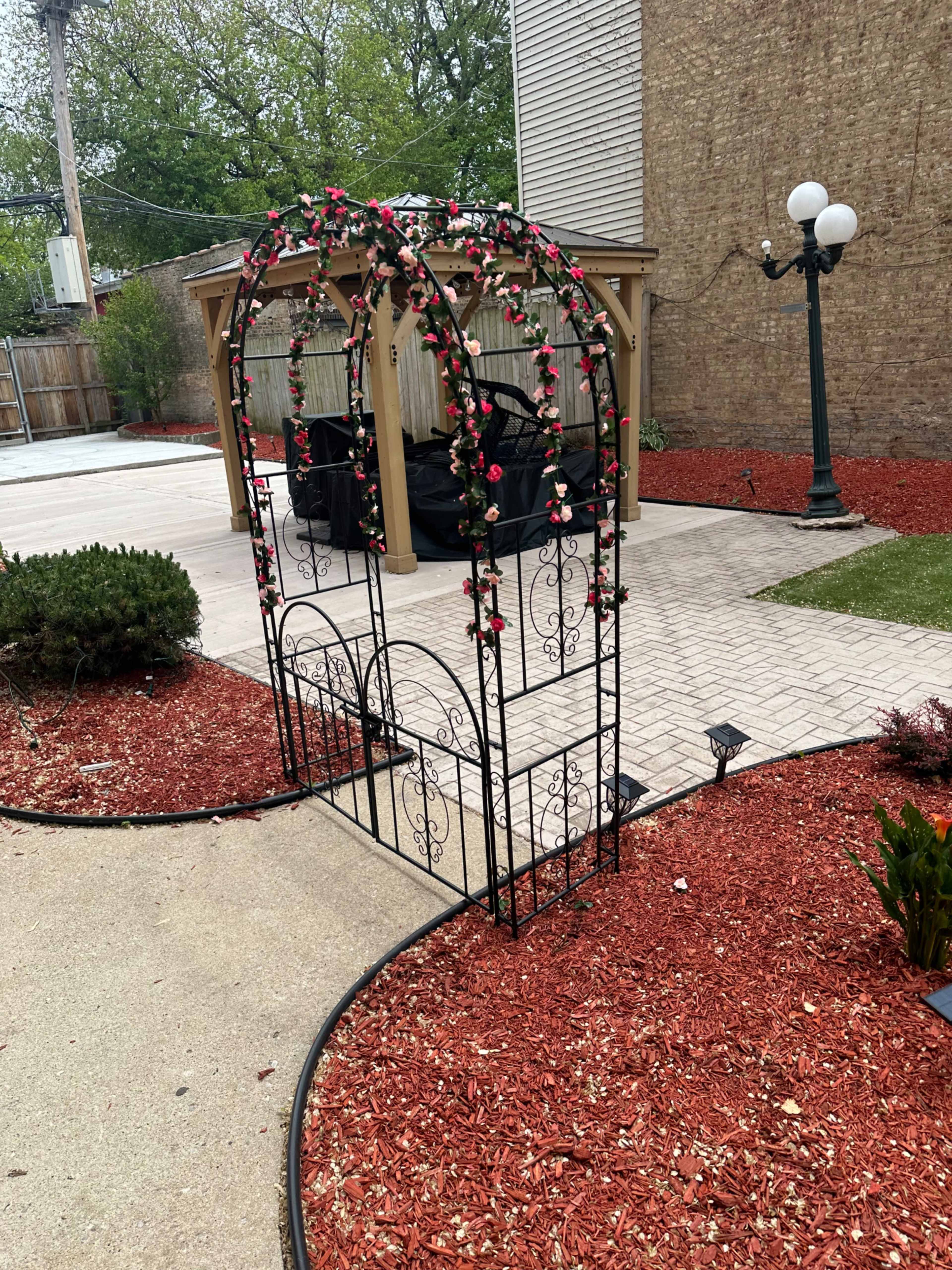 A black metal arch covered in artificial flowers, situated on a landscaped area with red mulch and surrounding greenery.