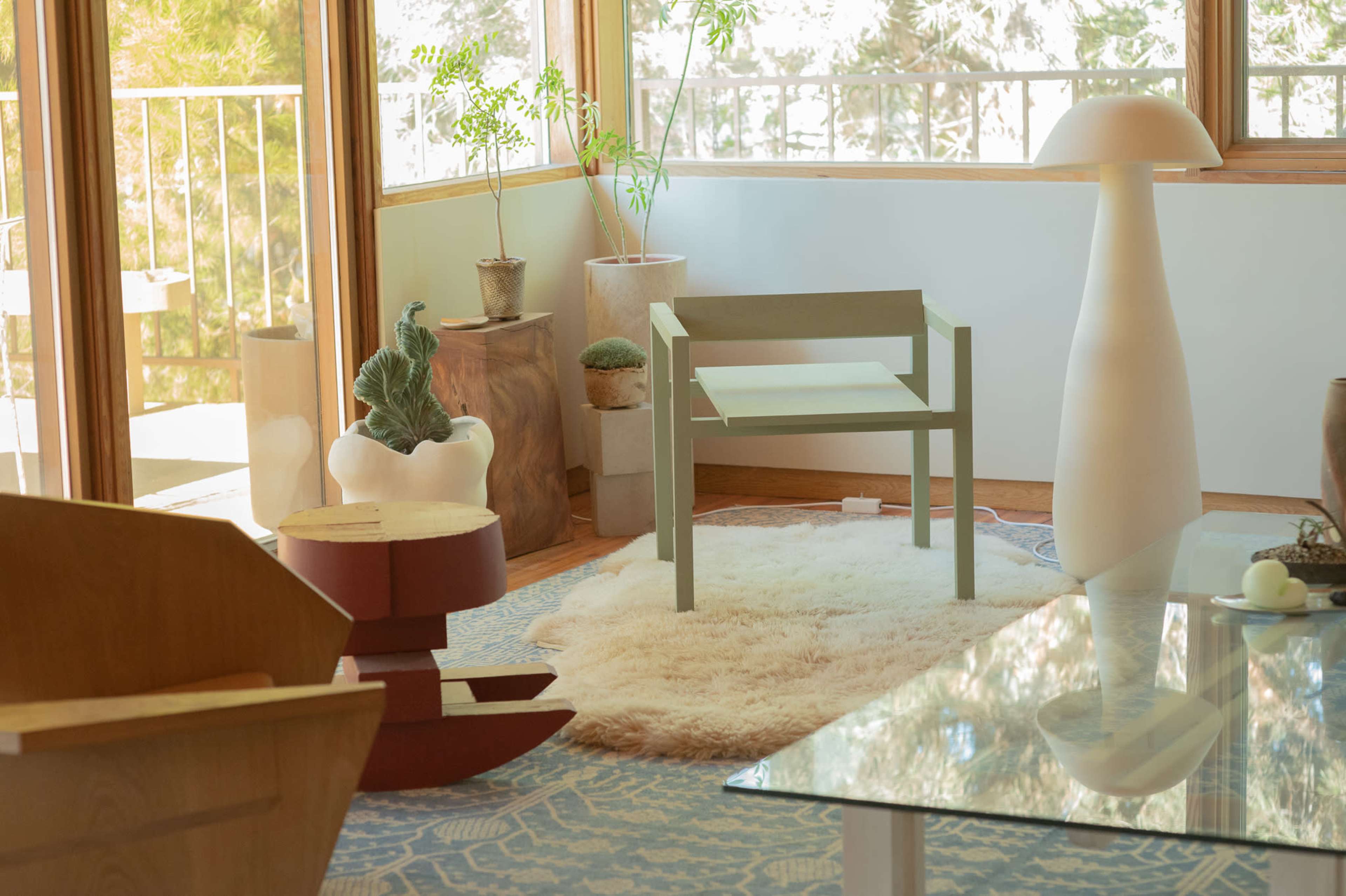 The scene features a modern living room with a green chair, a round rug, a glass coffee table, and various plants surrounded by large windows.