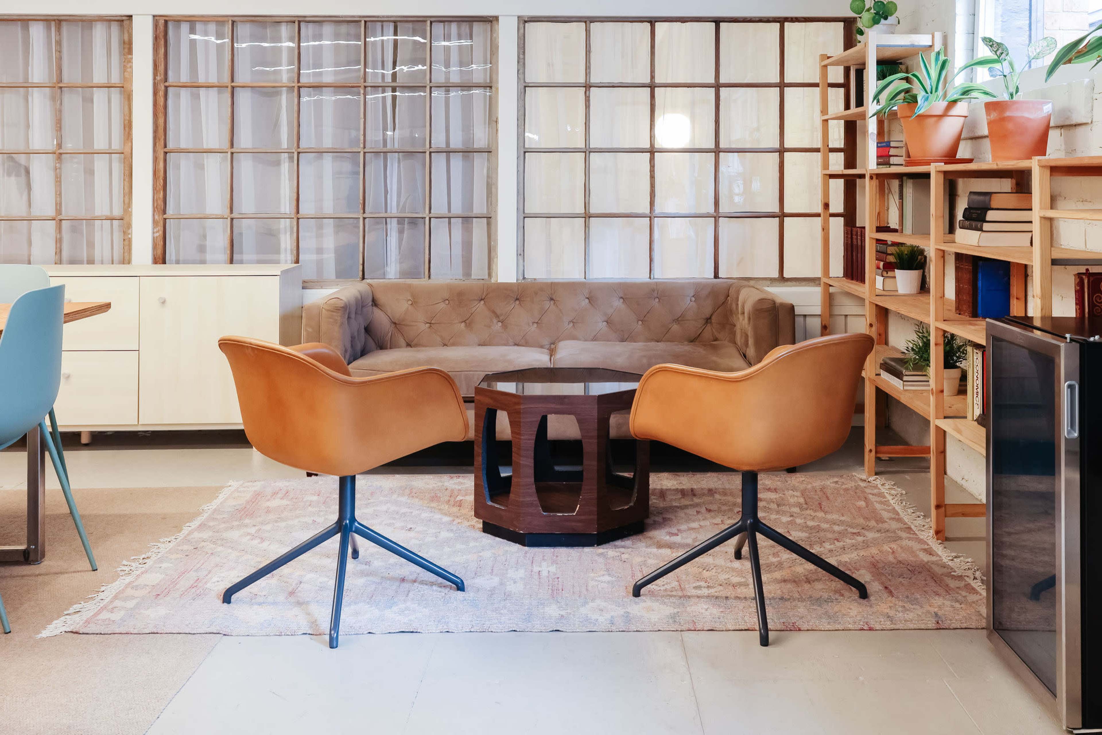 A well-furnished office space features a tufted sofa, two modern chairs, a wooden coffee table, and a bookshelf, with large windows allowing natural light.
