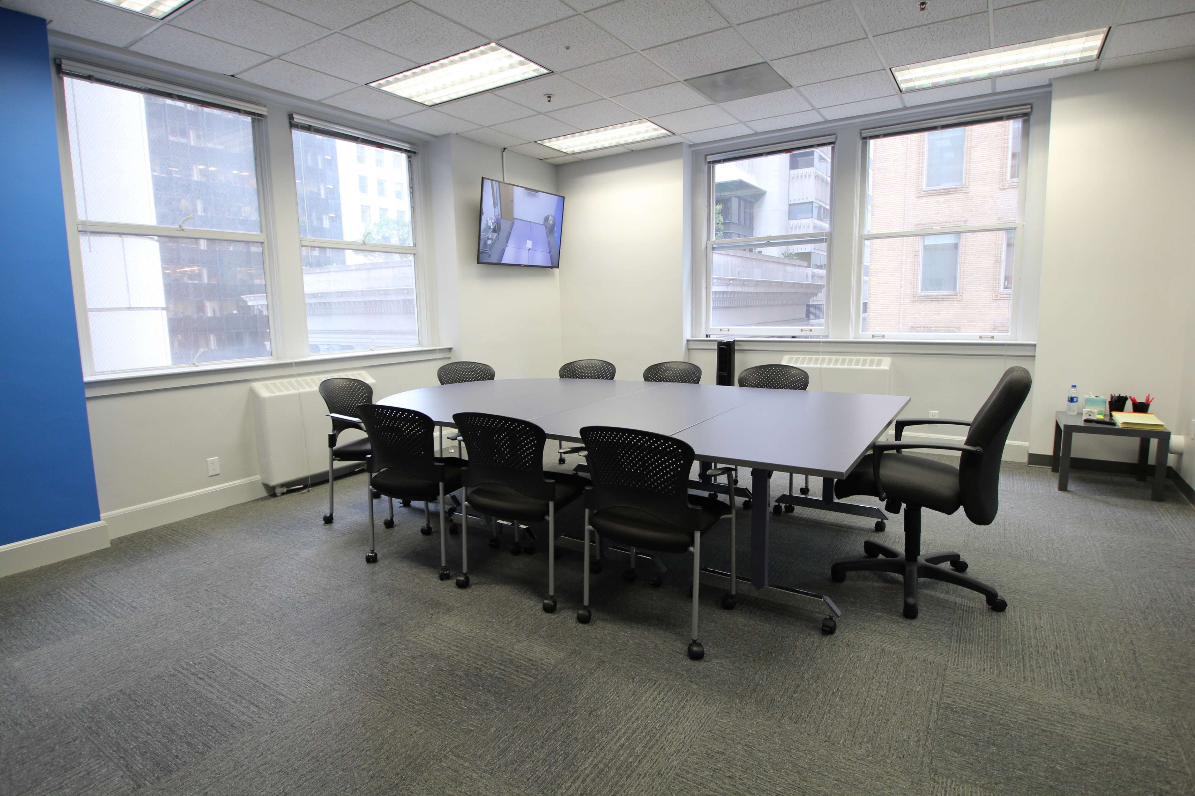A conference room with a large table surrounded by chairs, featuring a wall-mounted television and large windows.