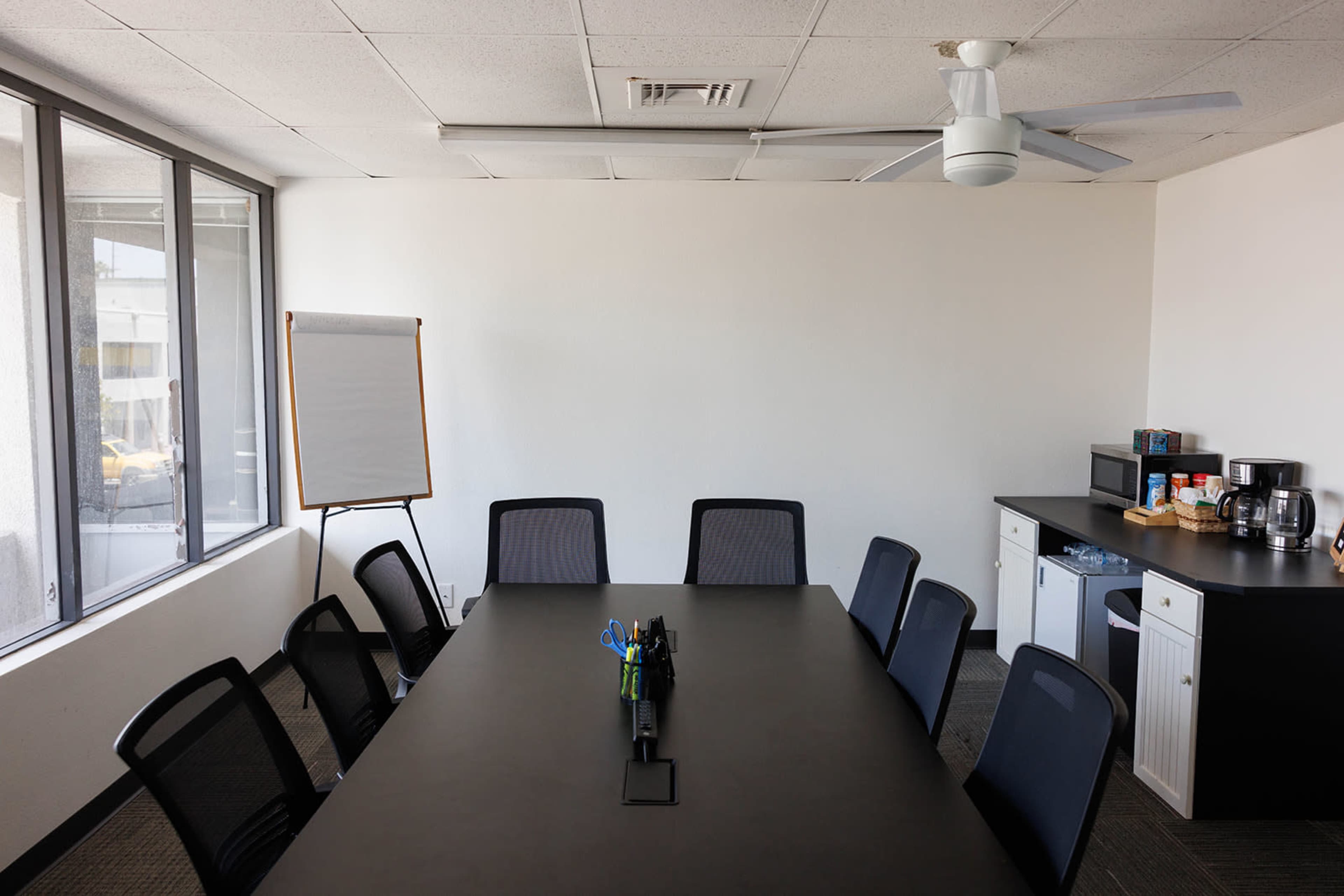 The image shows a conference room with a long black table surrounded by chairs, a whiteboard on an easel, and a kitchenette area with a coffee maker and snacks.