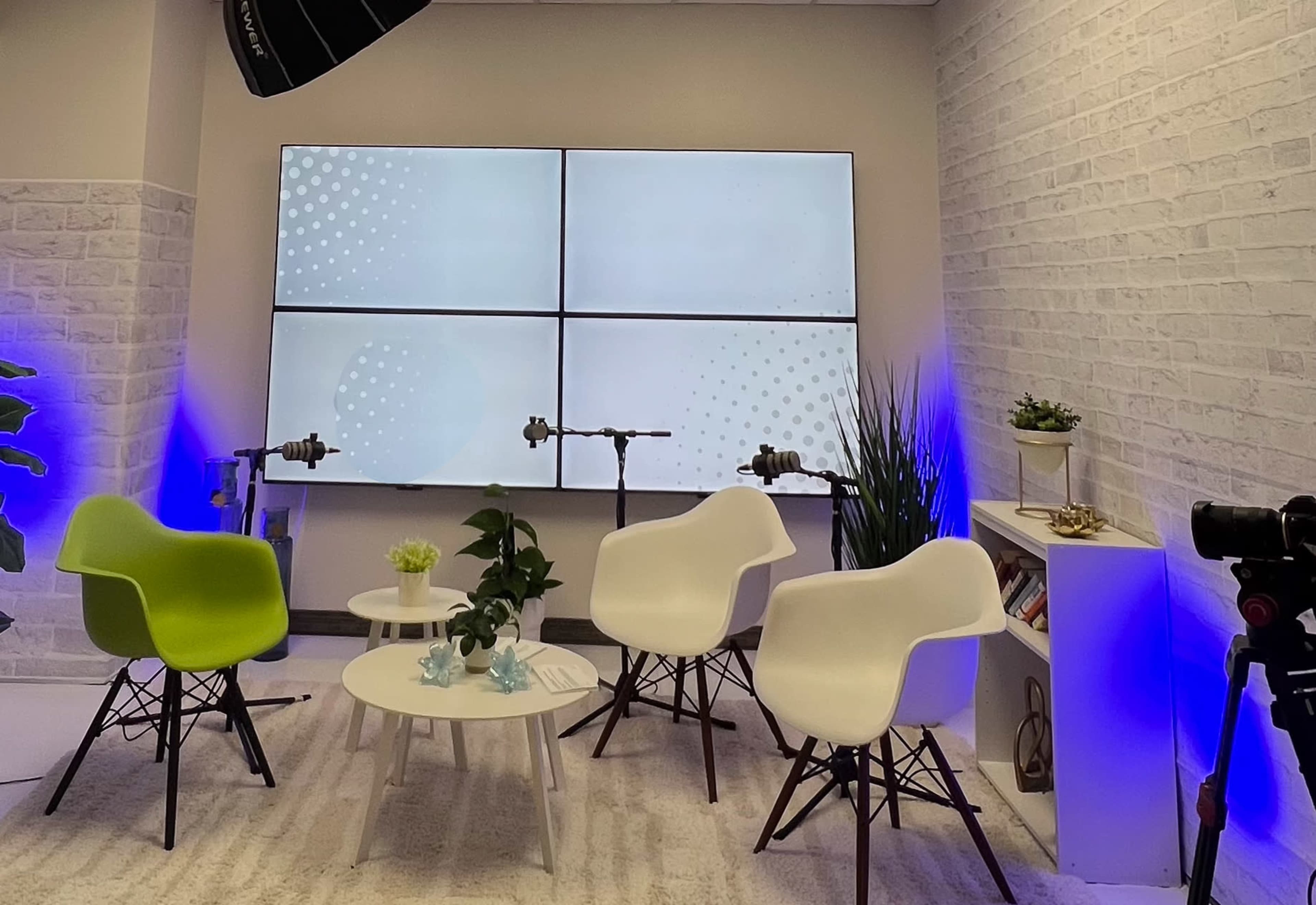The image shows a modern studio setup with four white chairs surrounding a round table, flanked by a large screen divided into four sections that displays a blank interface, and plants placed around the area.