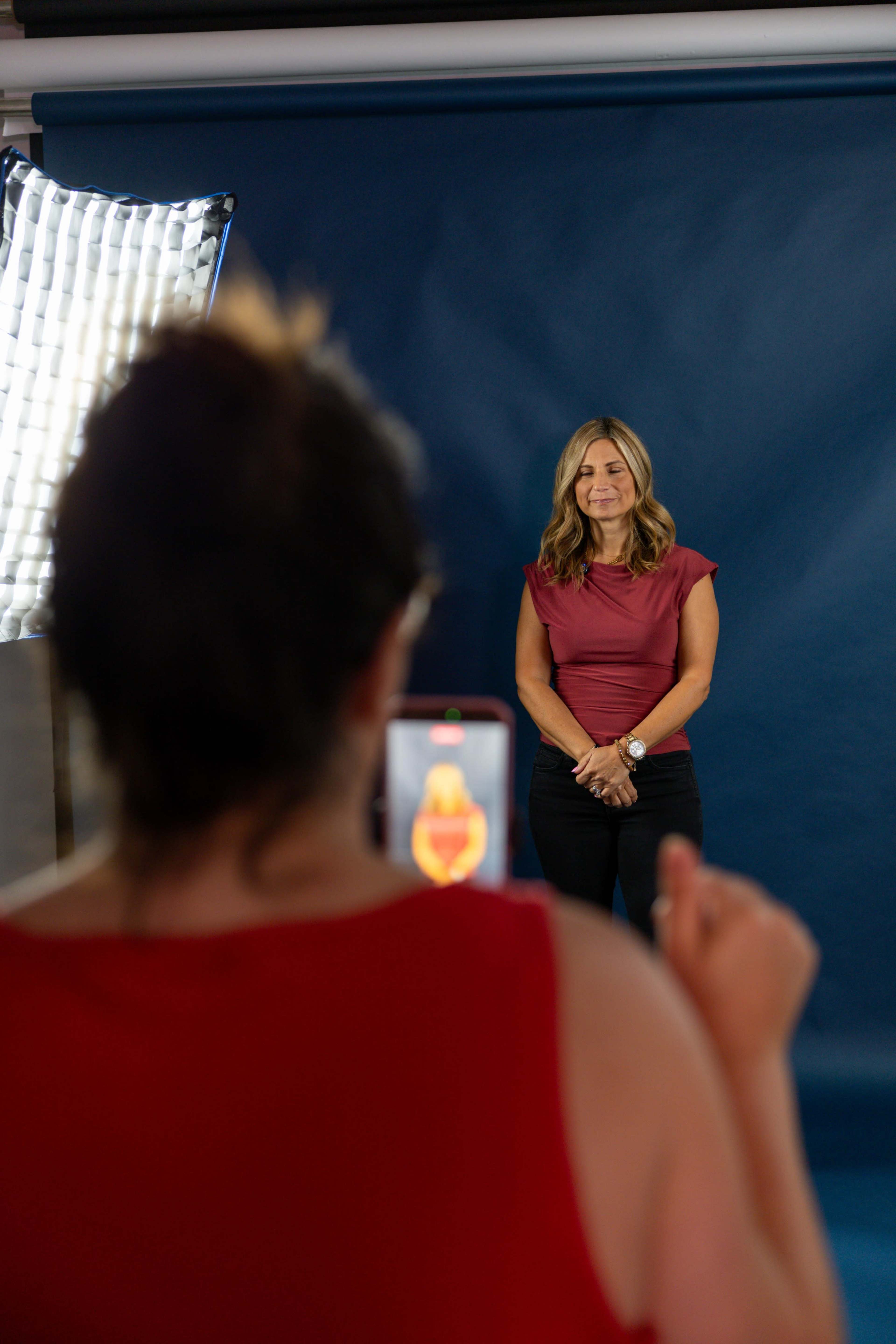 A woman is posing for a photo against a blue backdrop while another woman takes her picture with a smartphone.